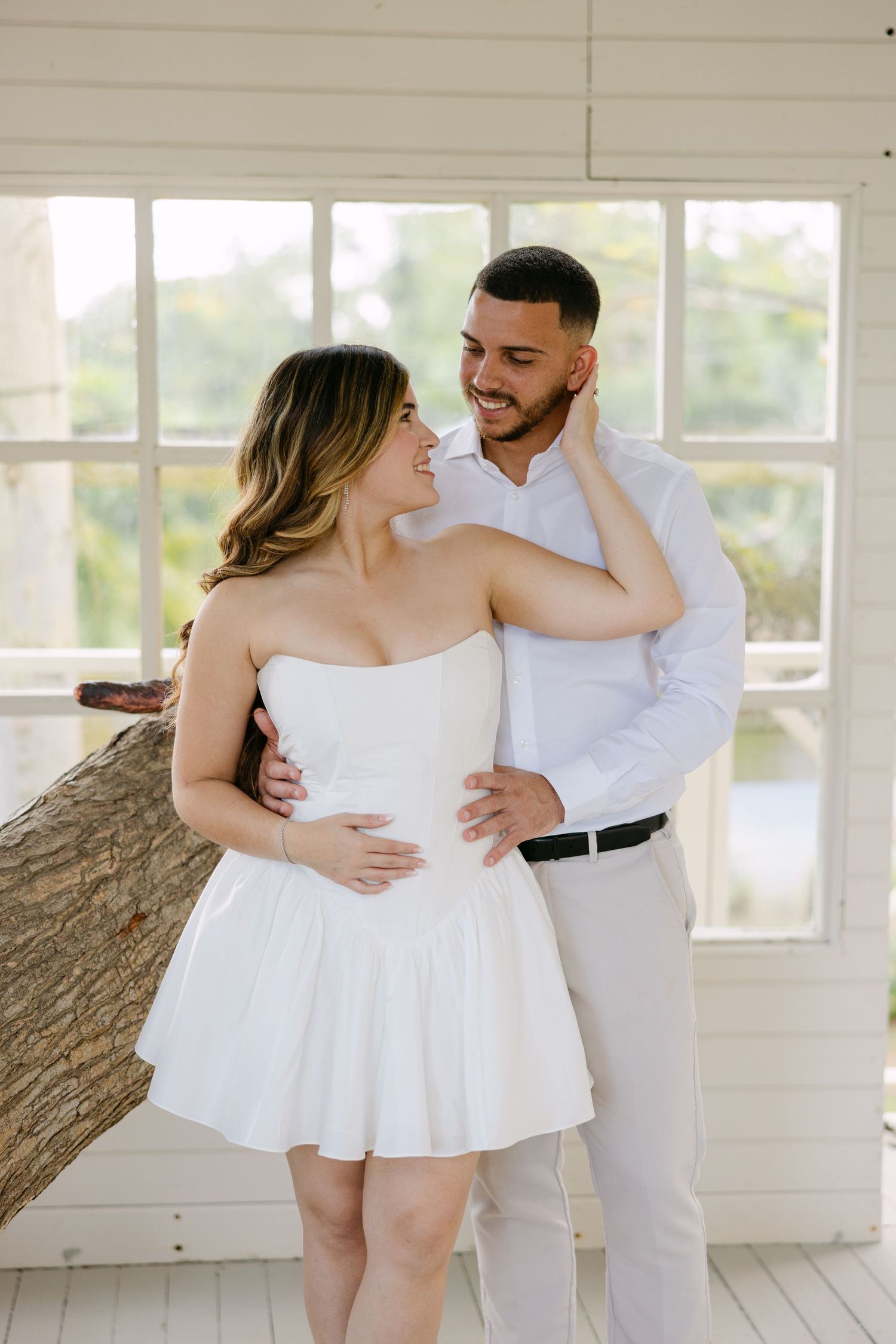 Romantic couple embraces in white attire during intimate indoor photoshoot with natural window lighting.