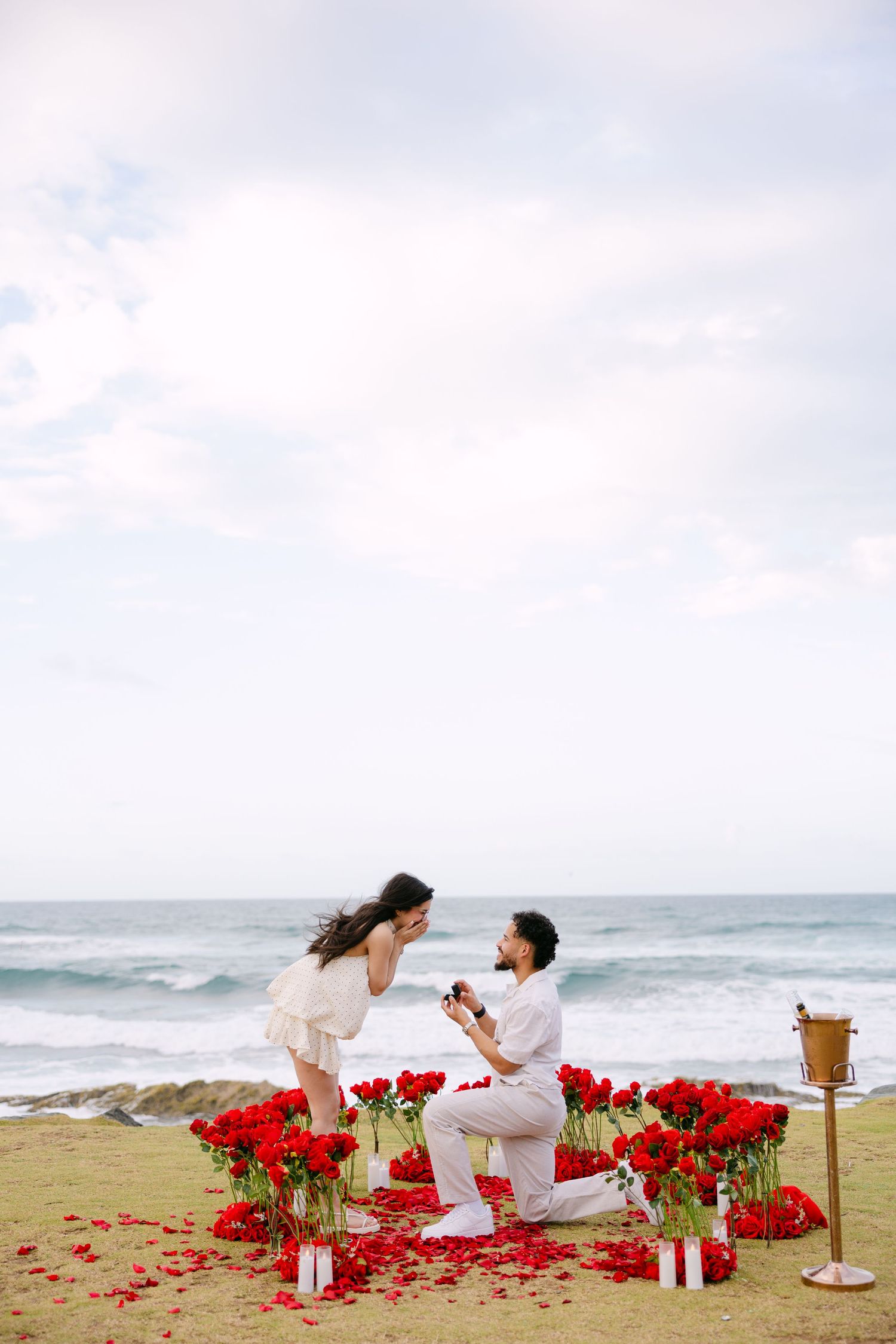 A romantic beach proposal setup with red rose petals and white decor elements along the ocean shoreline.