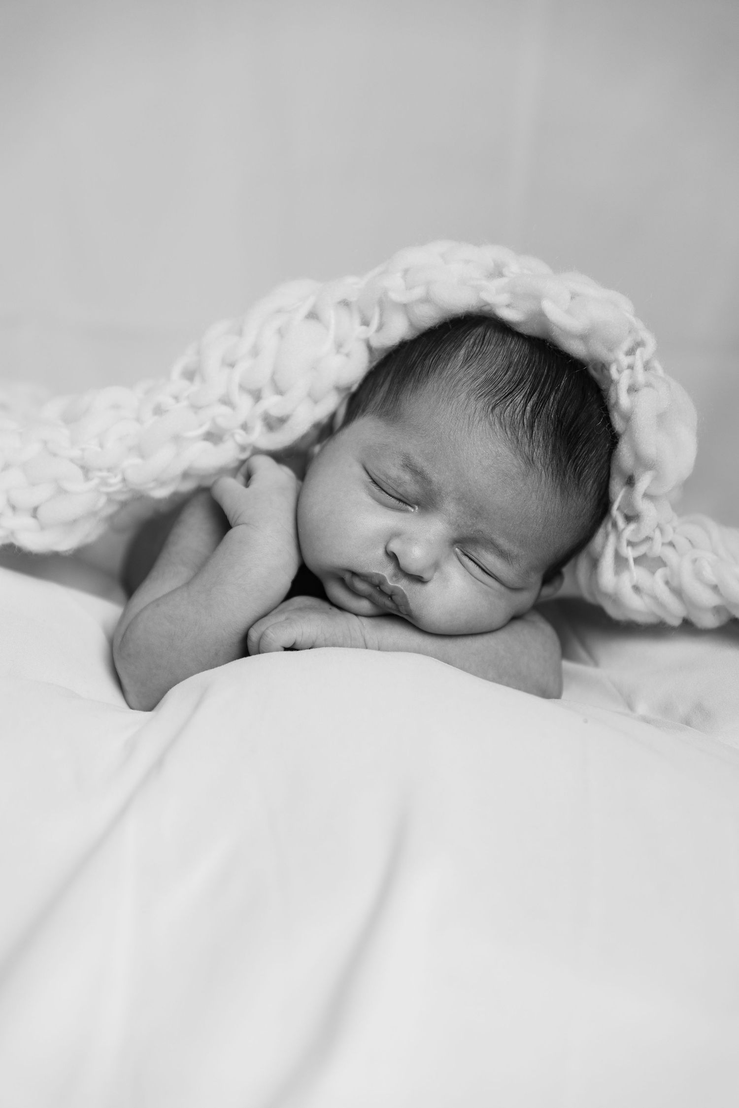 A sleepy newborn baby lies peacefully on white sheets in black and white tones.