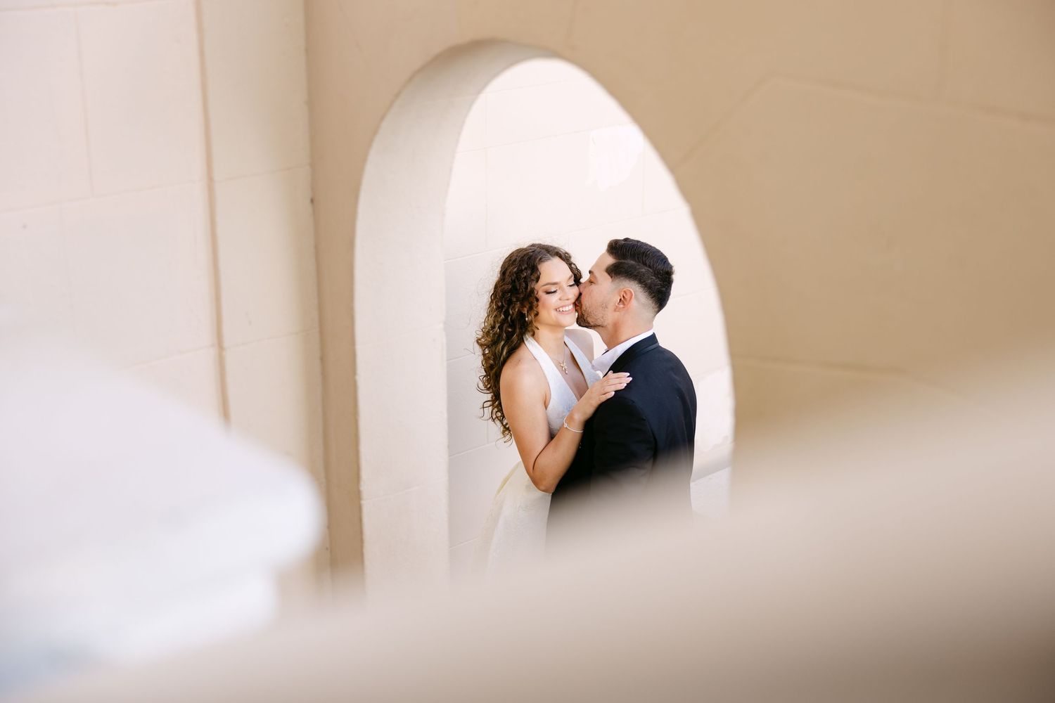 A romantic wedding couple shares an intimate kiss in a softly lit hallway with a neutral beige background.