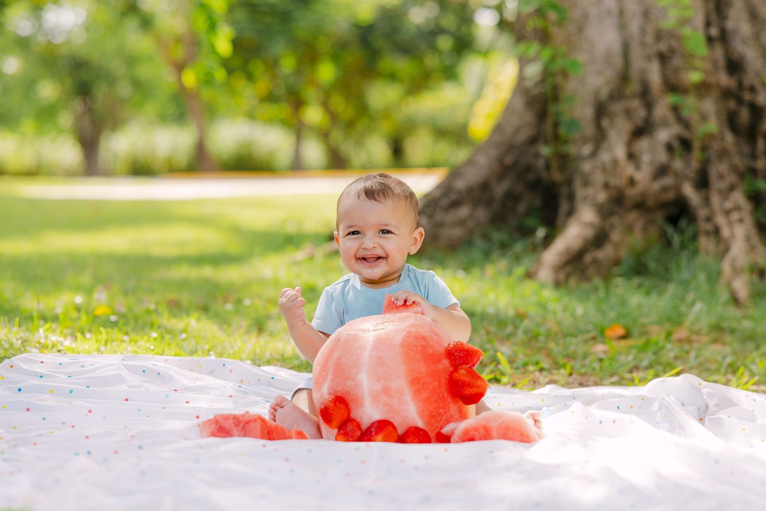 A small child in a blue shirt sits on a white blanket outdoors with a large red balloon.