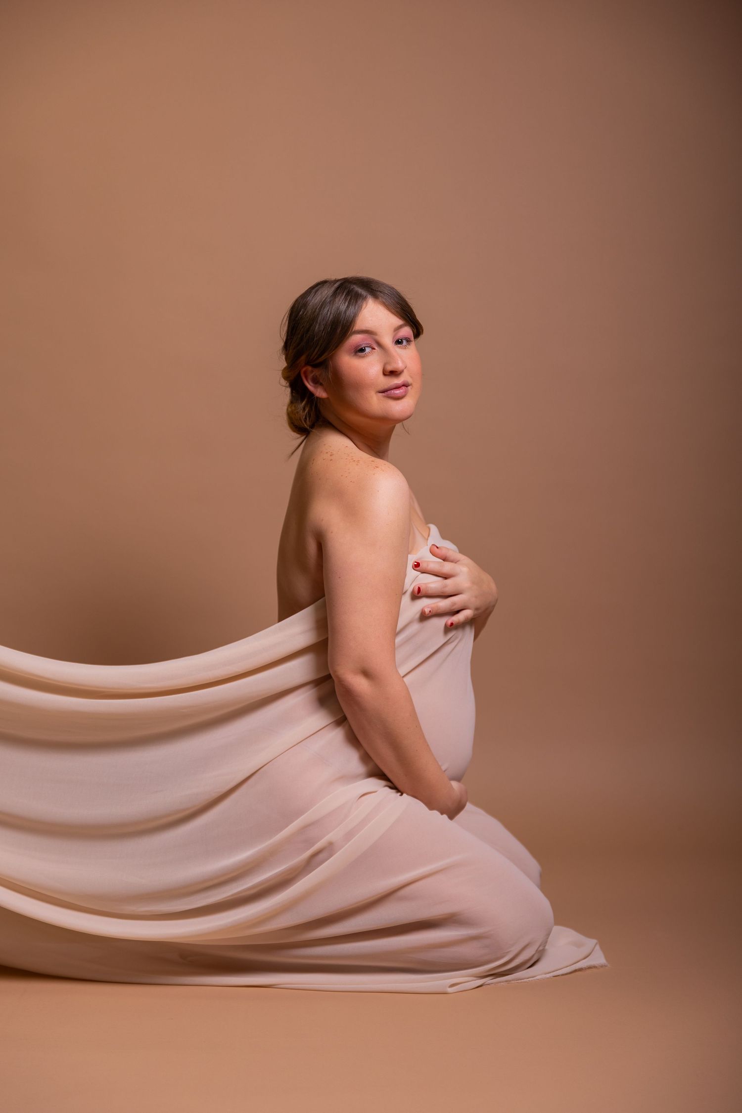 An artistic maternity portrait in soft blush tones featuring a person wrapped in flowing fabric against a neutral backdrop.