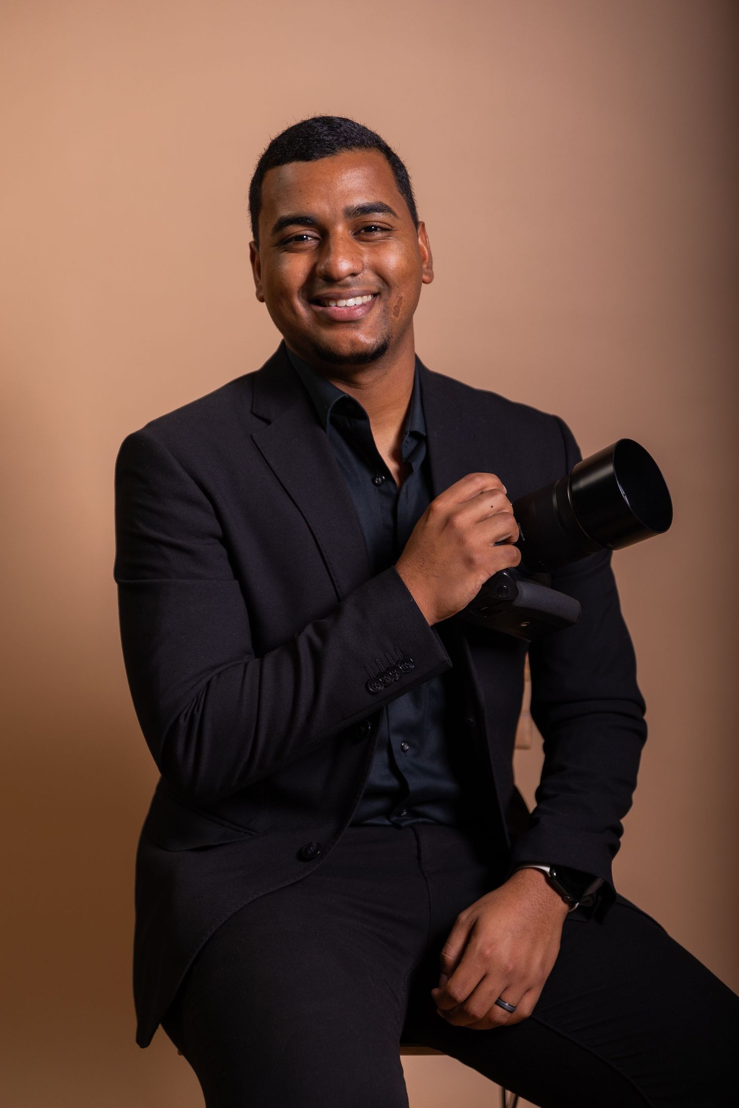Professional photographer in black suit holding camera and posing against neutral background.
