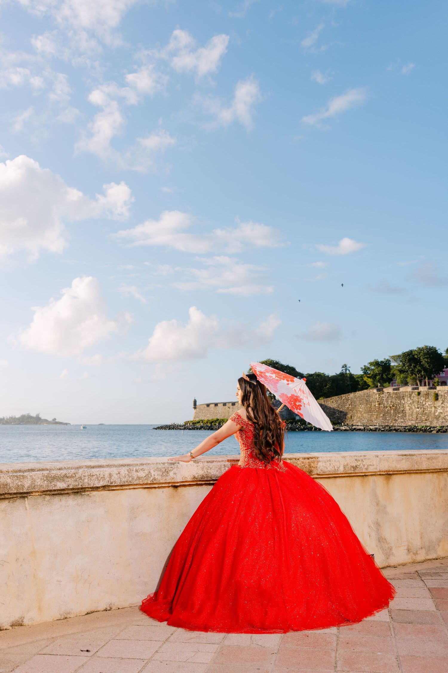 A series of photos showing a red ball gown against a seaside backdrop with stone walls and blue sky.