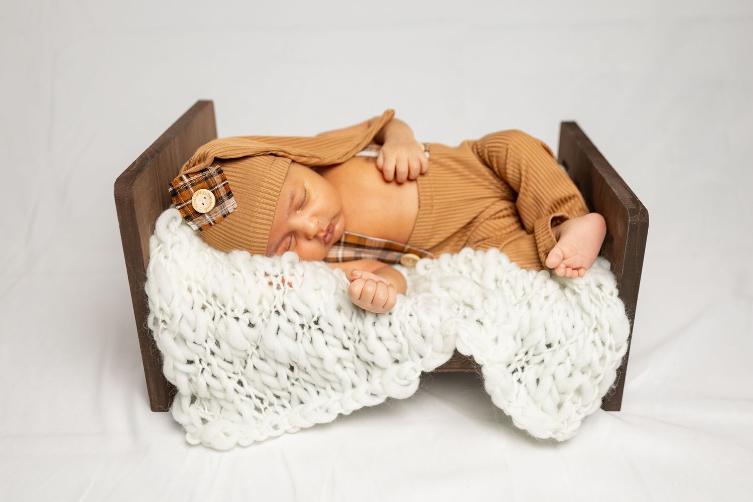 A baby lies wrapped in a white crocheted blanket on a brown pillow against a light background.