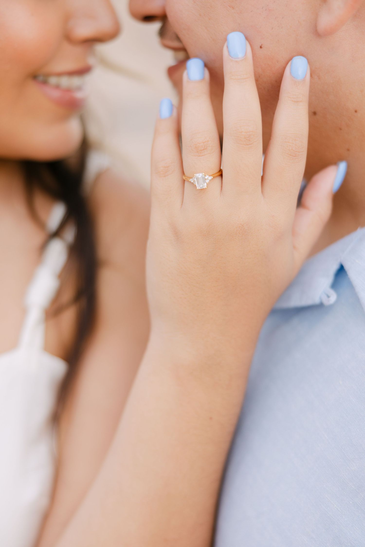 Close up view of hands with light blue nail polish showing delicate engagement ring against white backdrop.