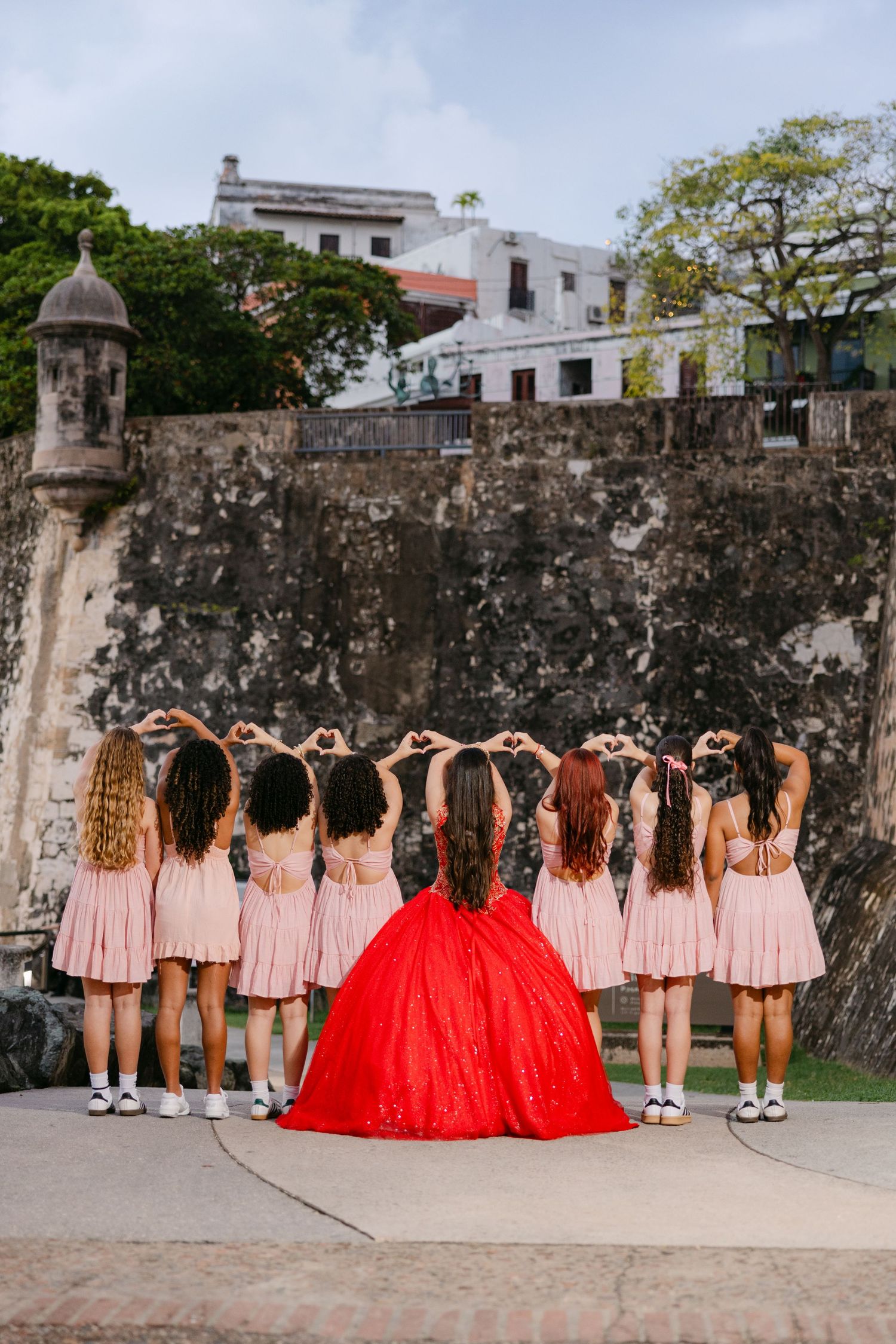 A group in matching pink dresses and one in a red gown pose with their backs turned against a historic stone wall.