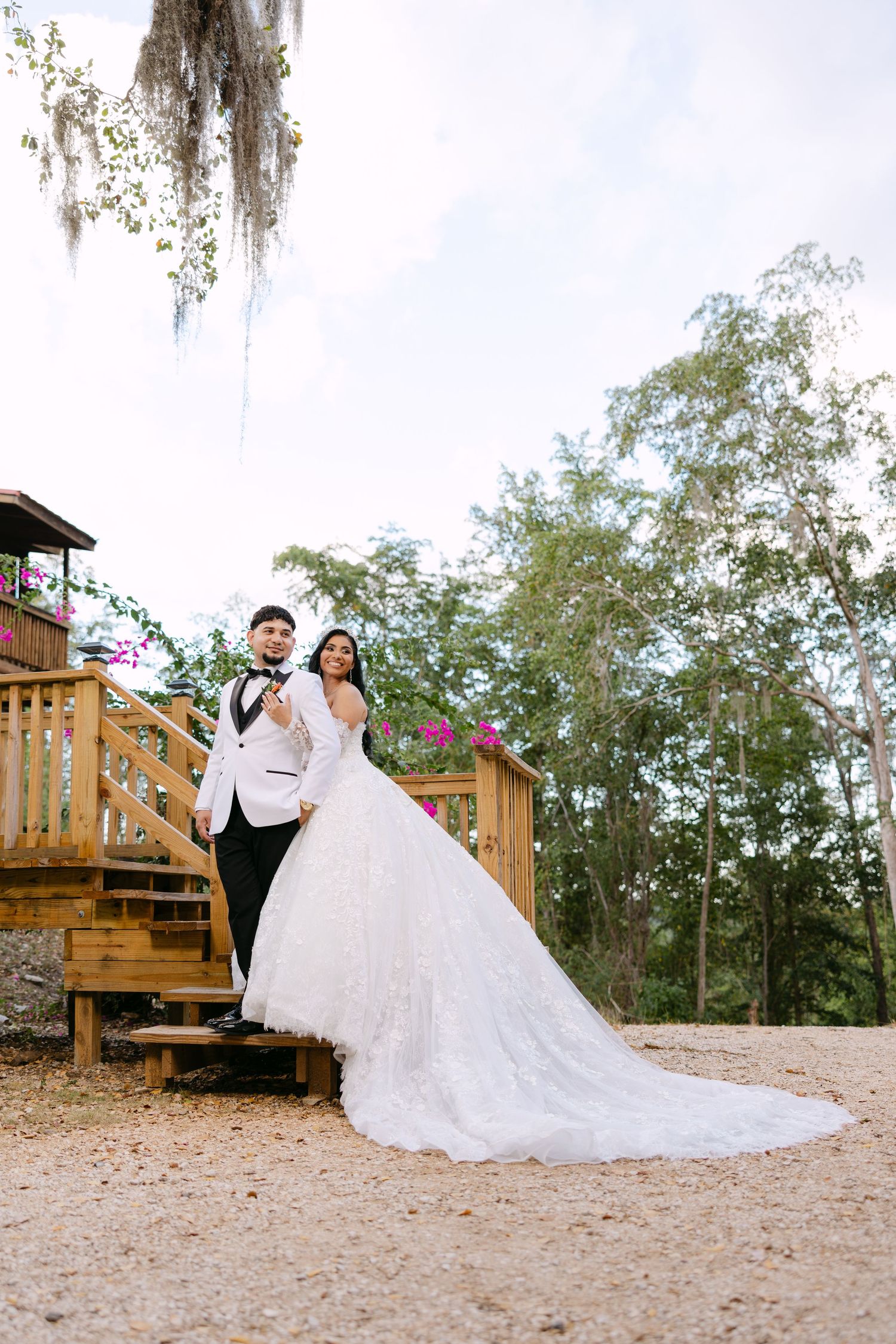 A newlywed couple poses on wooden stairs outdoors with Spanish moss and trees in the background.