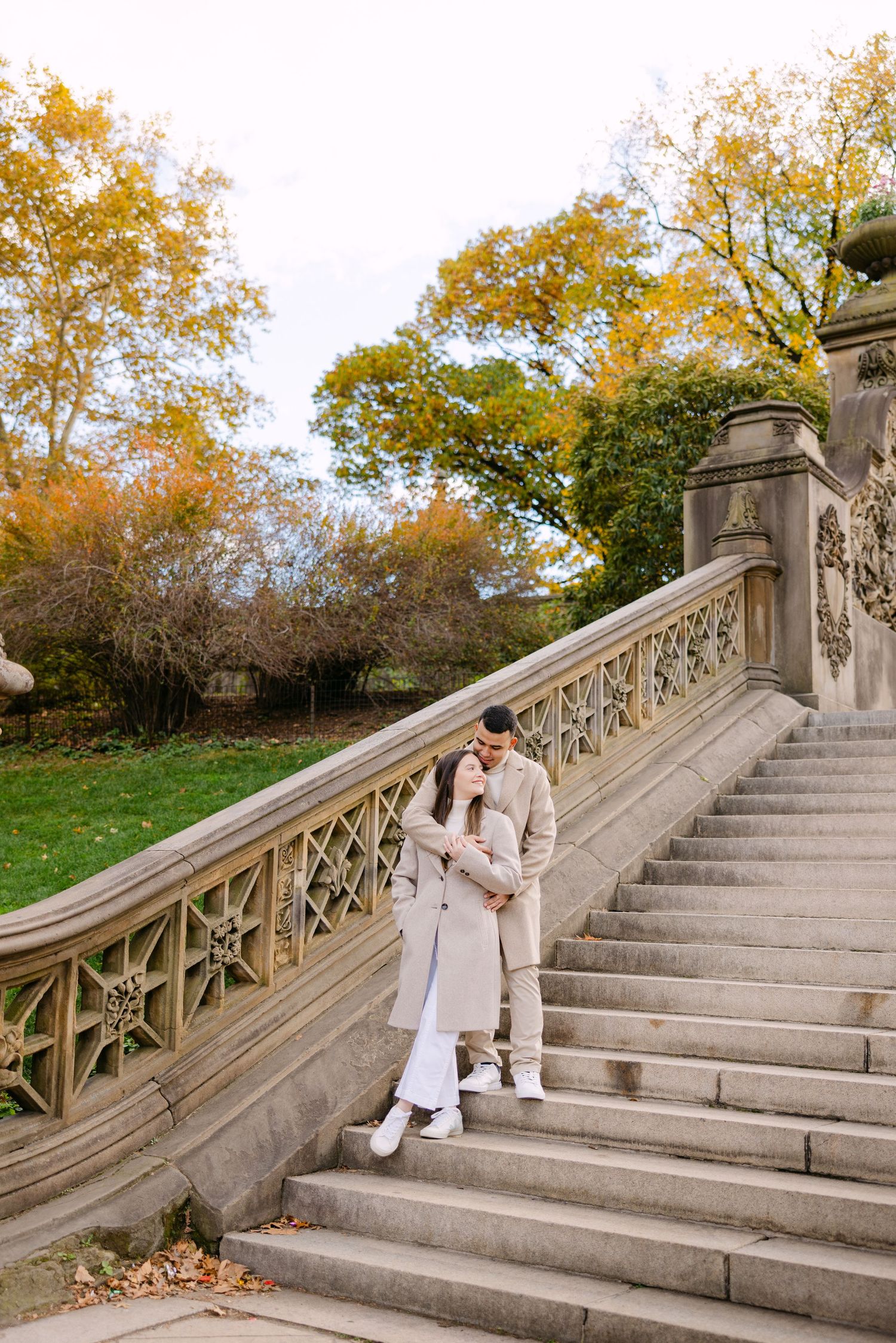 Two people in white outfits pose on stone steps surrounded by autumn trees with golden foliage.