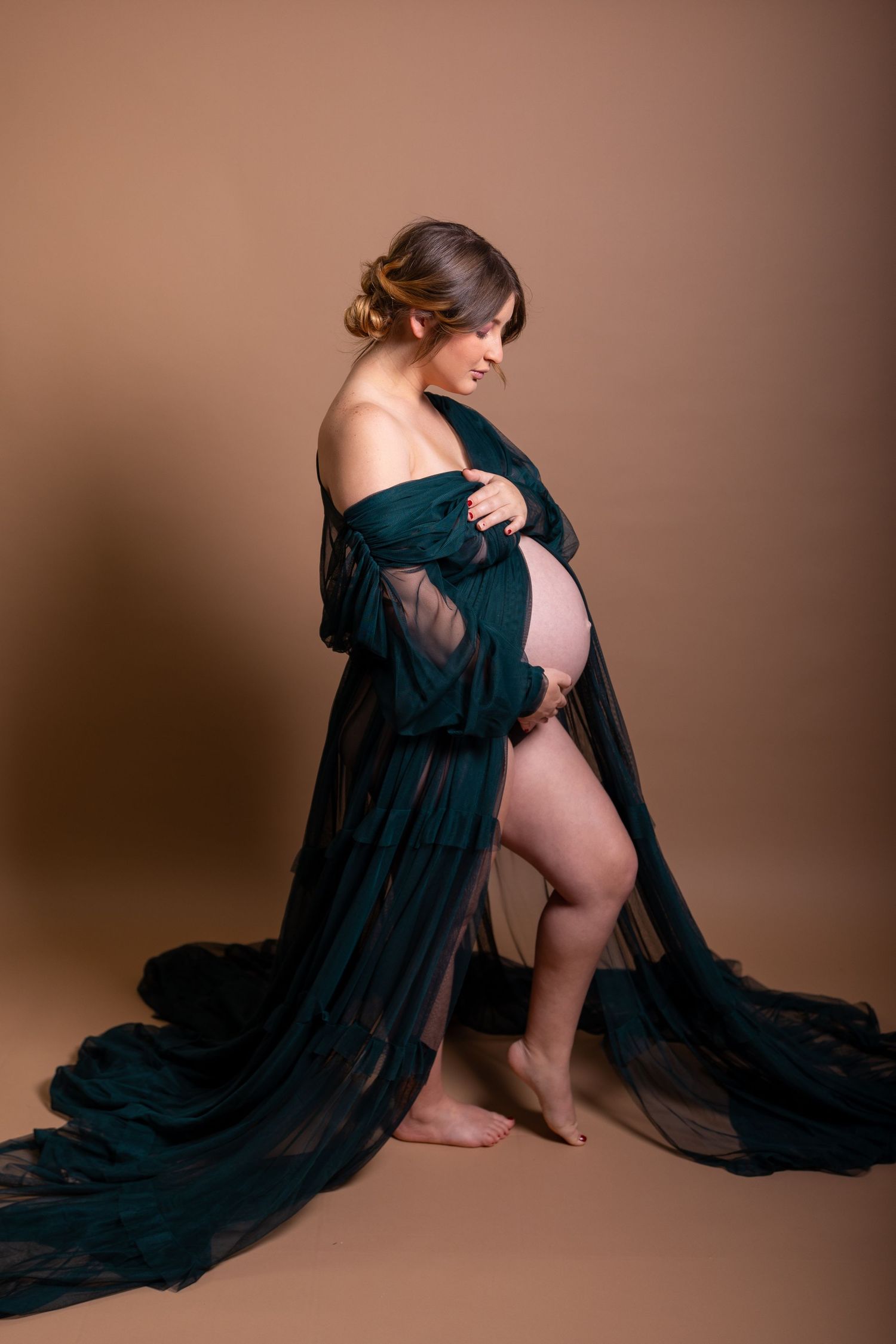 A pregnant woman poses in a flowing dark teal robe against a brown backdrop during an artistic maternity photoshoot.