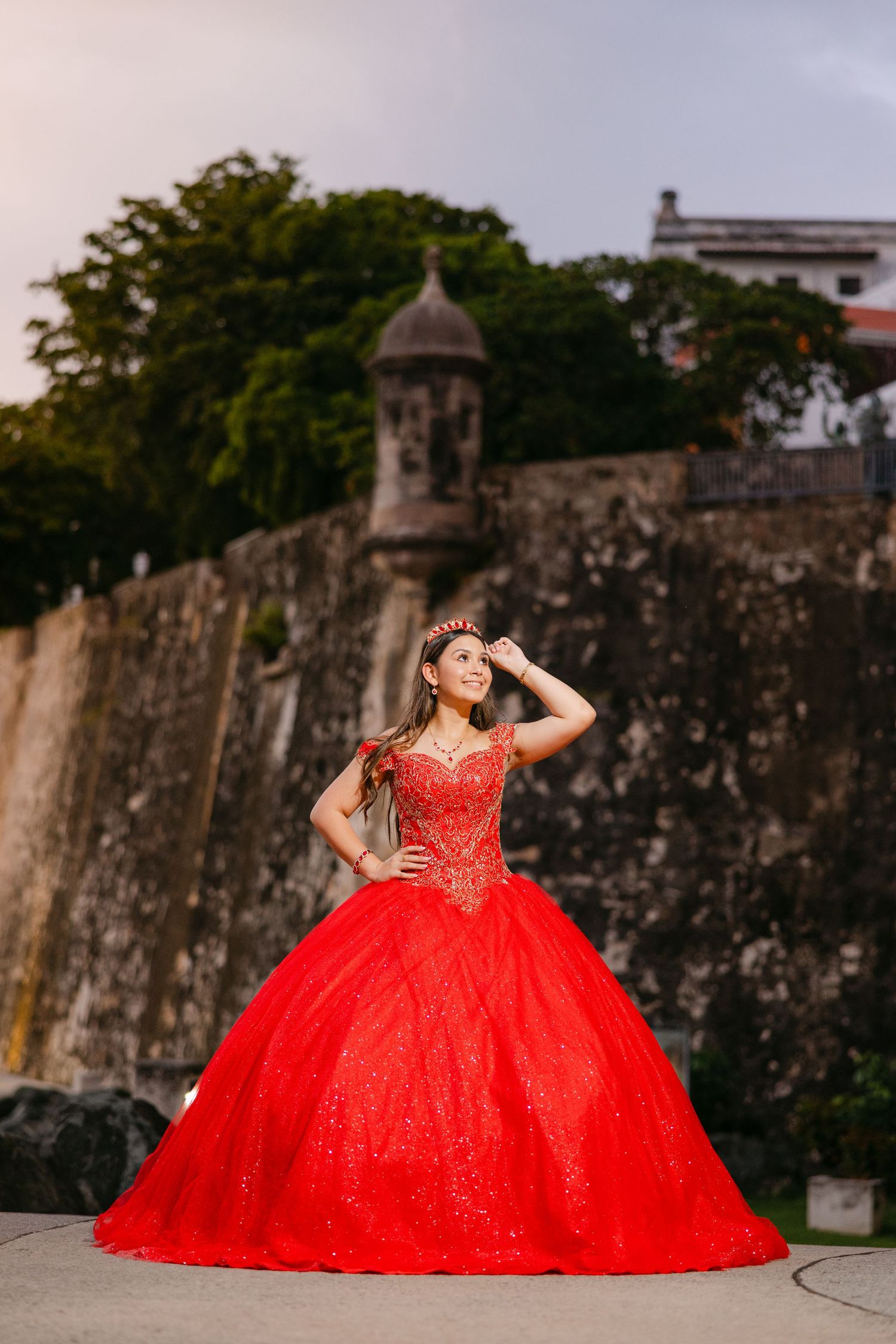 A stunning red quinceañera ball gown photographed against historic stone fortress walls in dramatic poses.