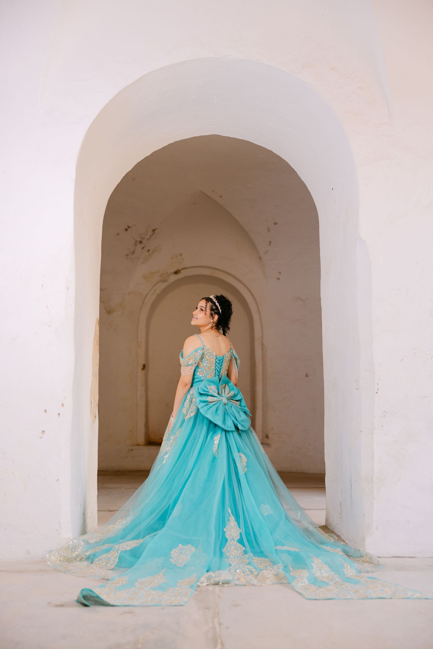 A stunning turquoise ball gown with a dramatic bow and flowing train photographed in an elegant arched doorway.