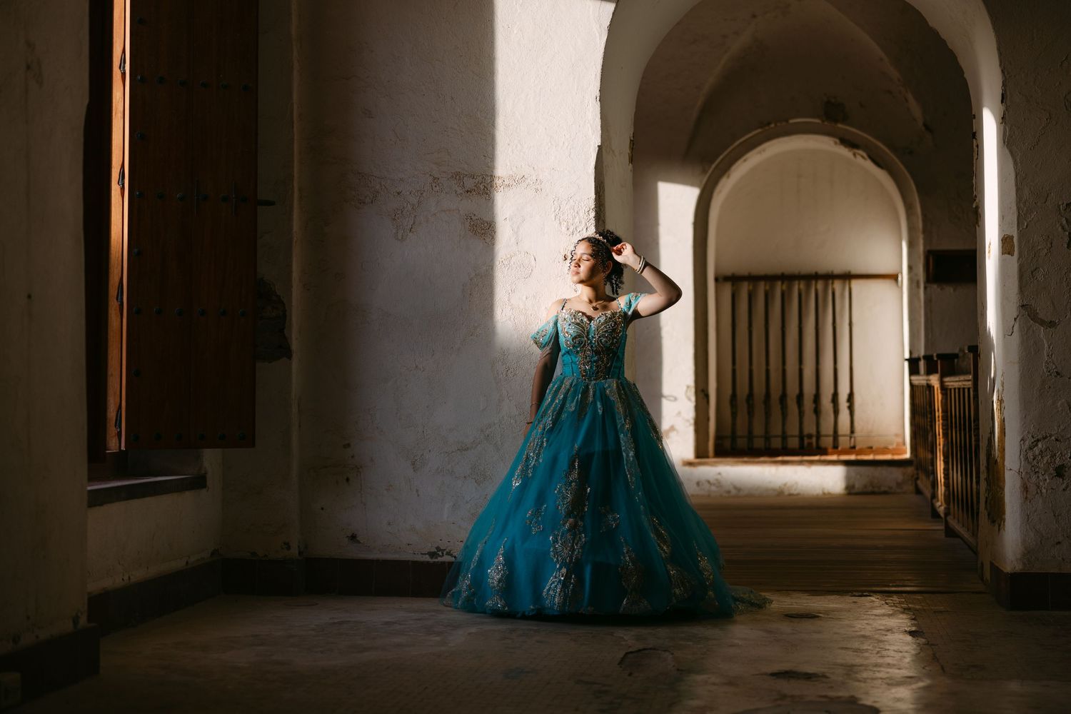 A person in a teal ballgown poses dramatically in an arched stone hallway with dramatic lighting.