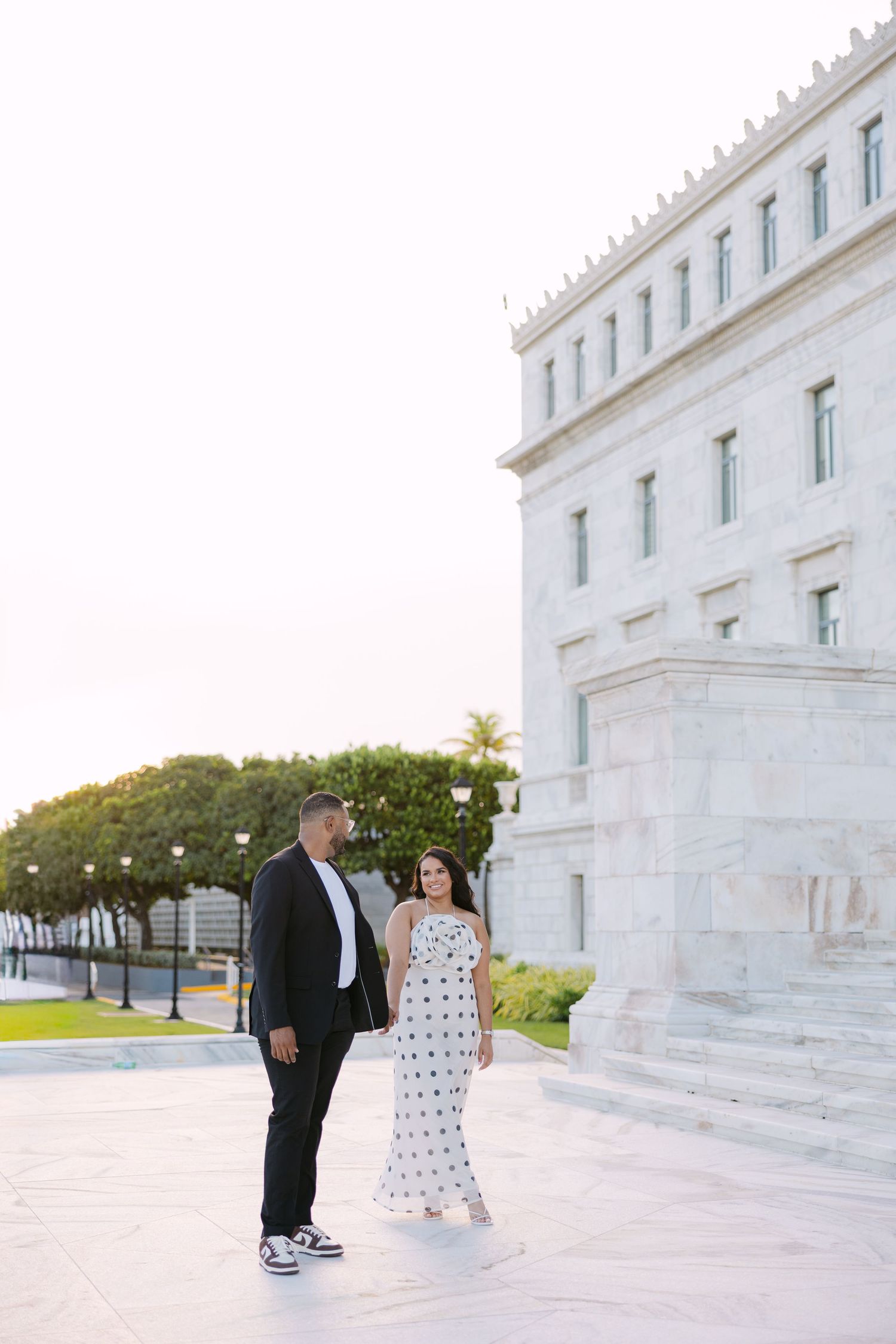A couple poses in formal attire against a white classical building exterior during a seaside photoshoot.