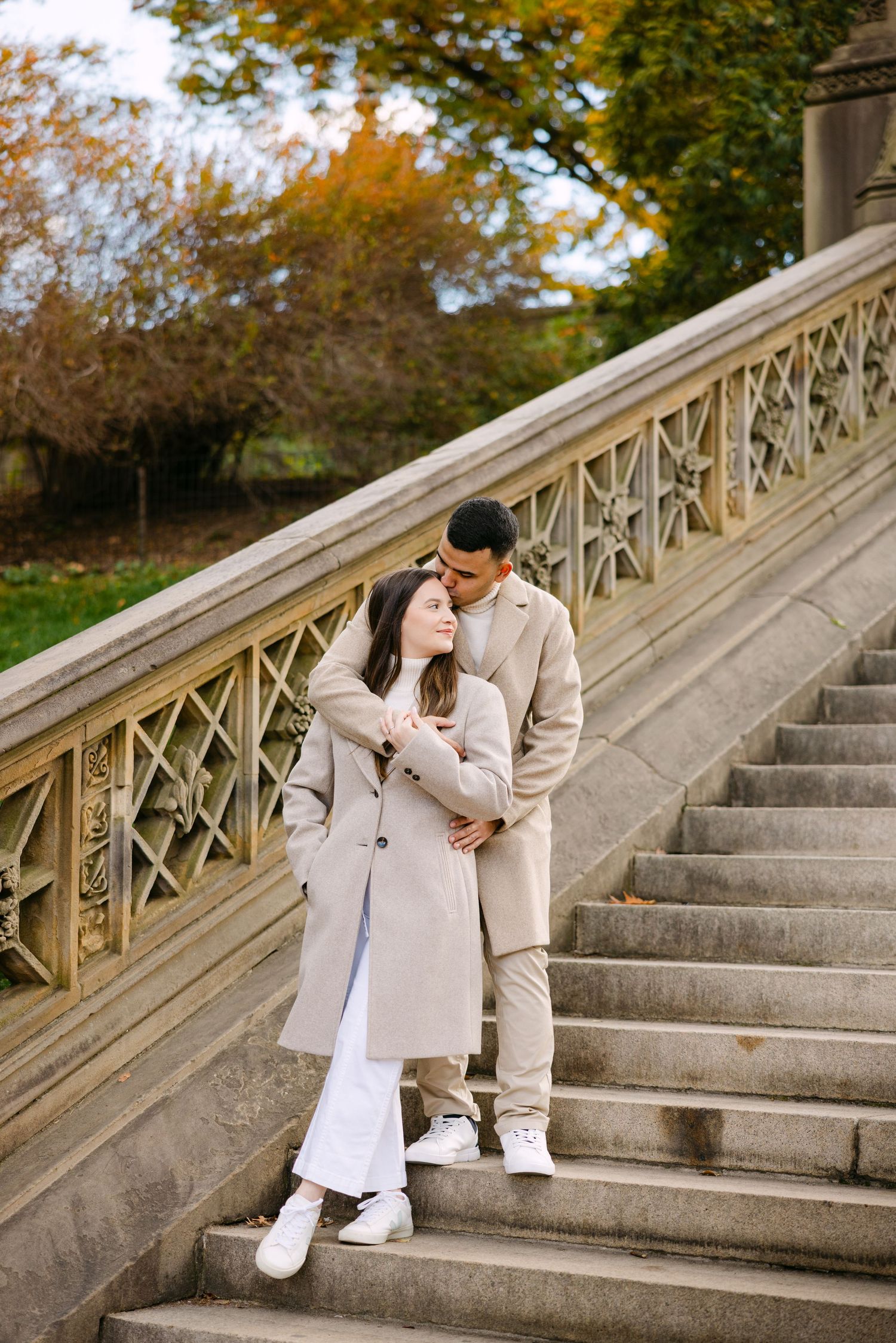 A couple sits together on stone steps surrounded by autumn foliage while sharing a romantic moment.