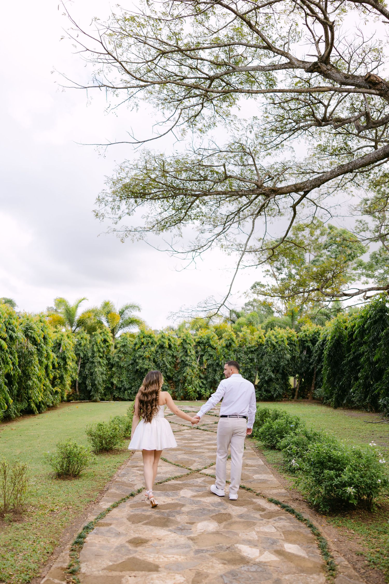 A couple walks hand in hand along a scenic garden path bordered by lush greenery and palm trees.