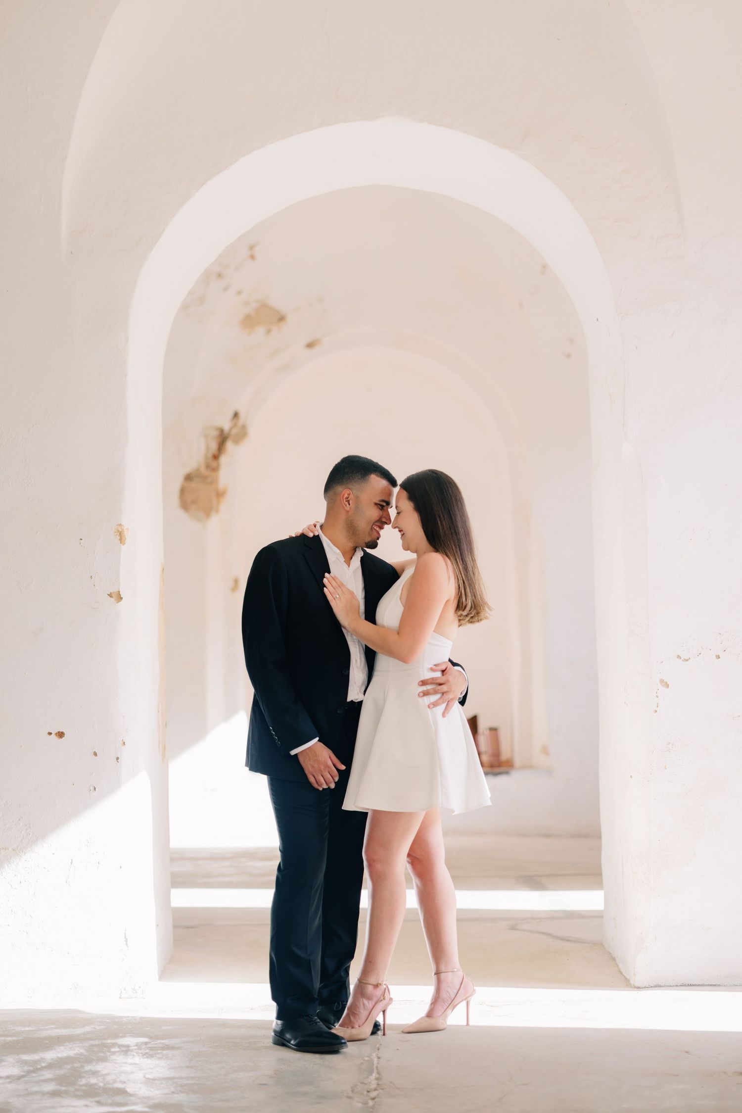 Romantic couple embraces in an elegant white archway, dressed in formal black suit and short white dress with heels.