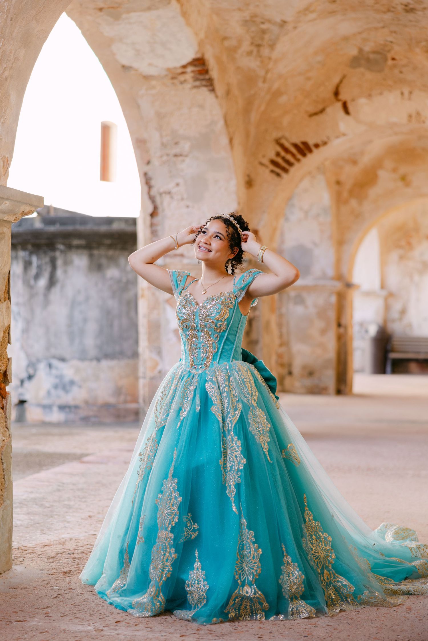 A person in a flowing turquoise and gold ball gown poses gracefully in an ancient stone archway.