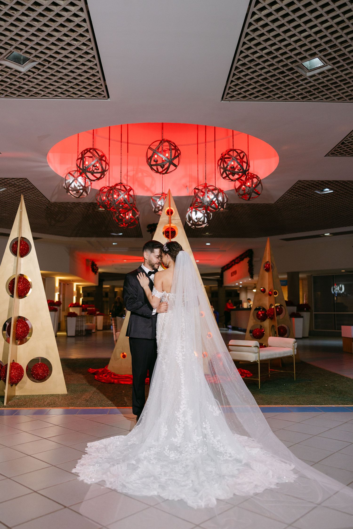 A couple stands beneath red geometric pendant lights and decorative Christmas trees in an elegant reception venue.