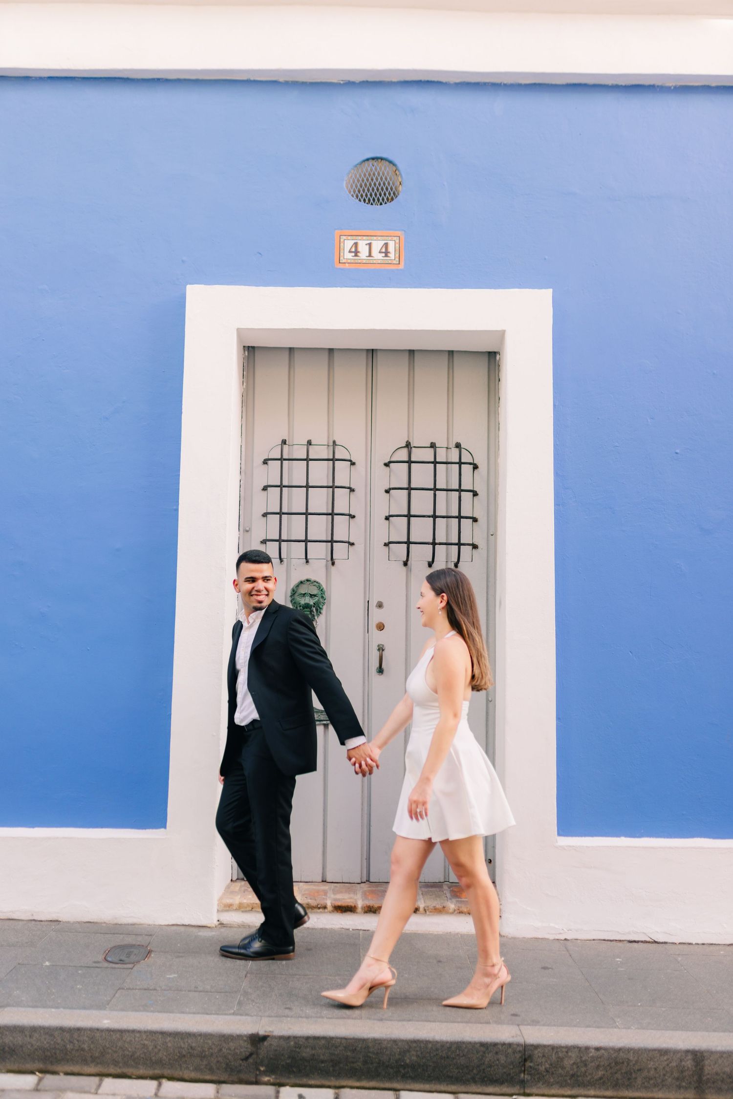 A couple stands in front of a gray door on a bright blue building, dressed elegantly in black and white.