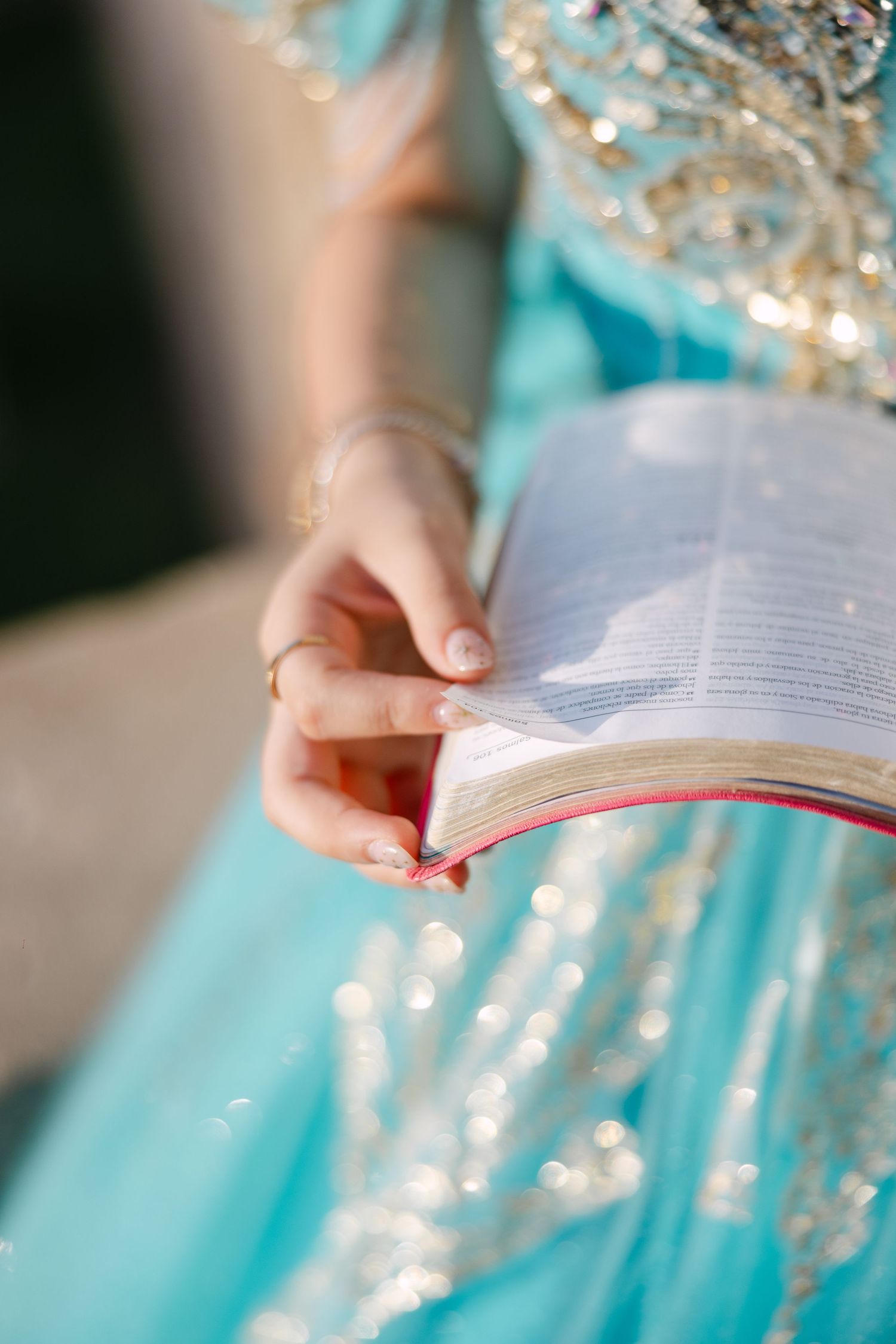 Hands hold an open book against a turquoise dress with sparkling details in soft natural lighting.