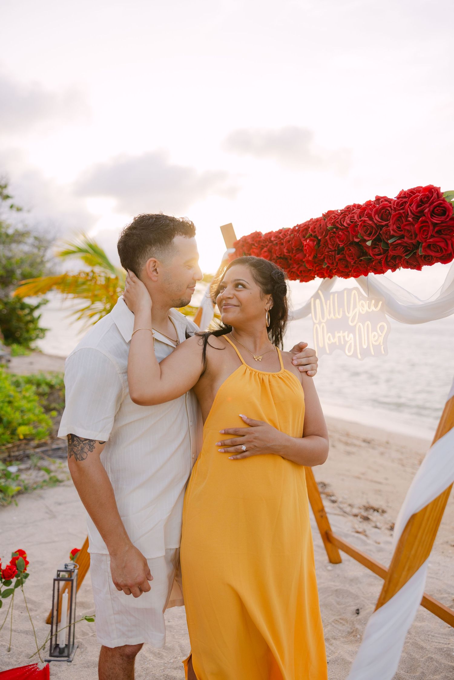 Two people share tender moments at sunset on a beach with red flowers and decorative lanterns in the background.