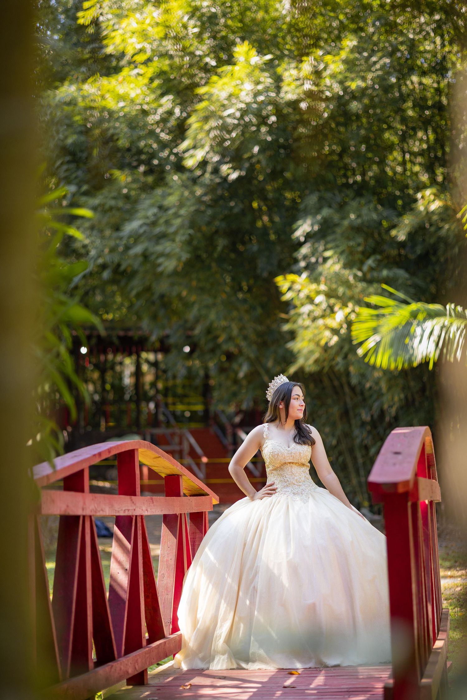 A bride in a ballgown wedding dress poses on a red bridge surrounded by lush greenery.