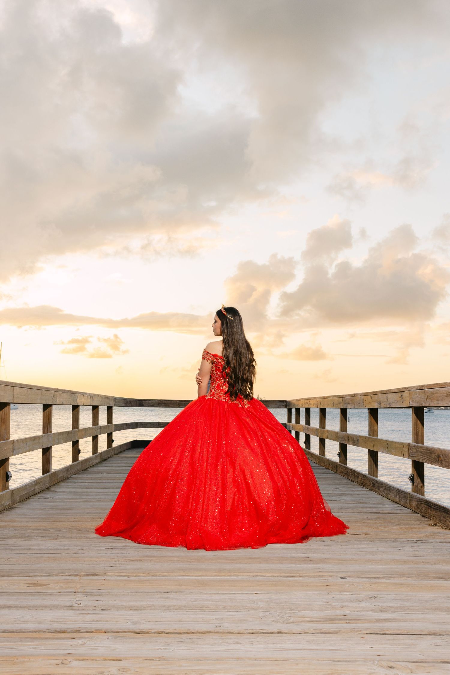 A flowing red ball gown creates a dramatic silhouette against a wooden pier at sunset with cloudy skies.