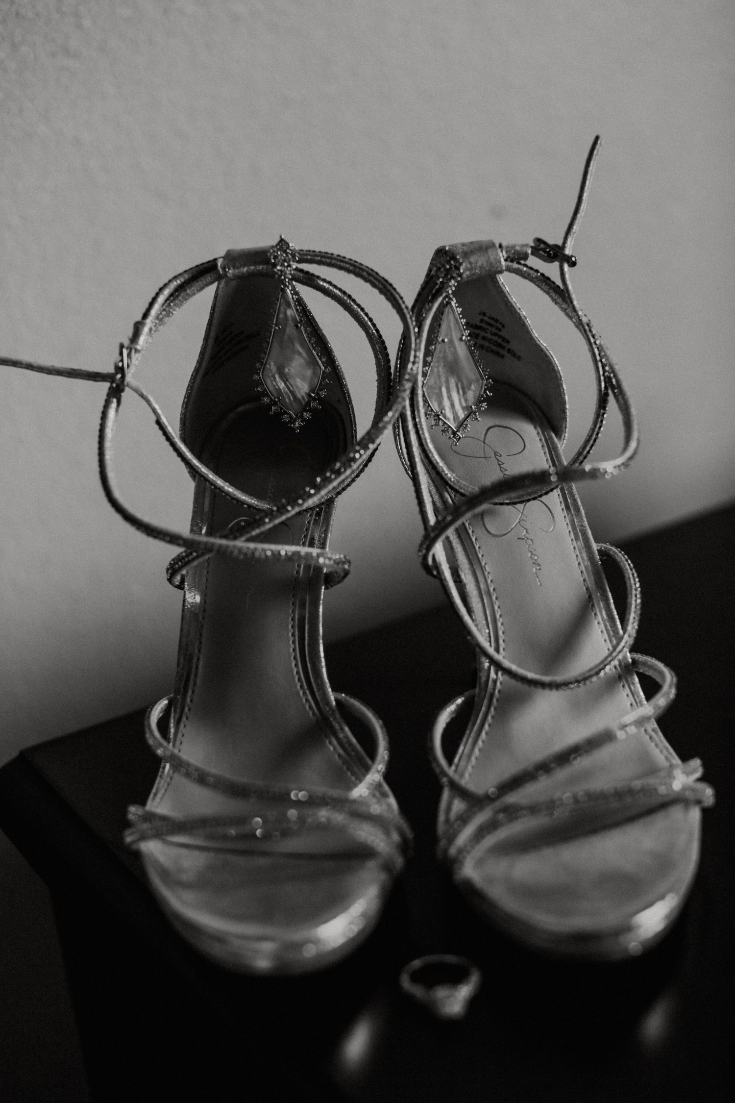 Black and white photo of strappy high-heel bridal sandals displayed on a dark surface with a ring nearby.