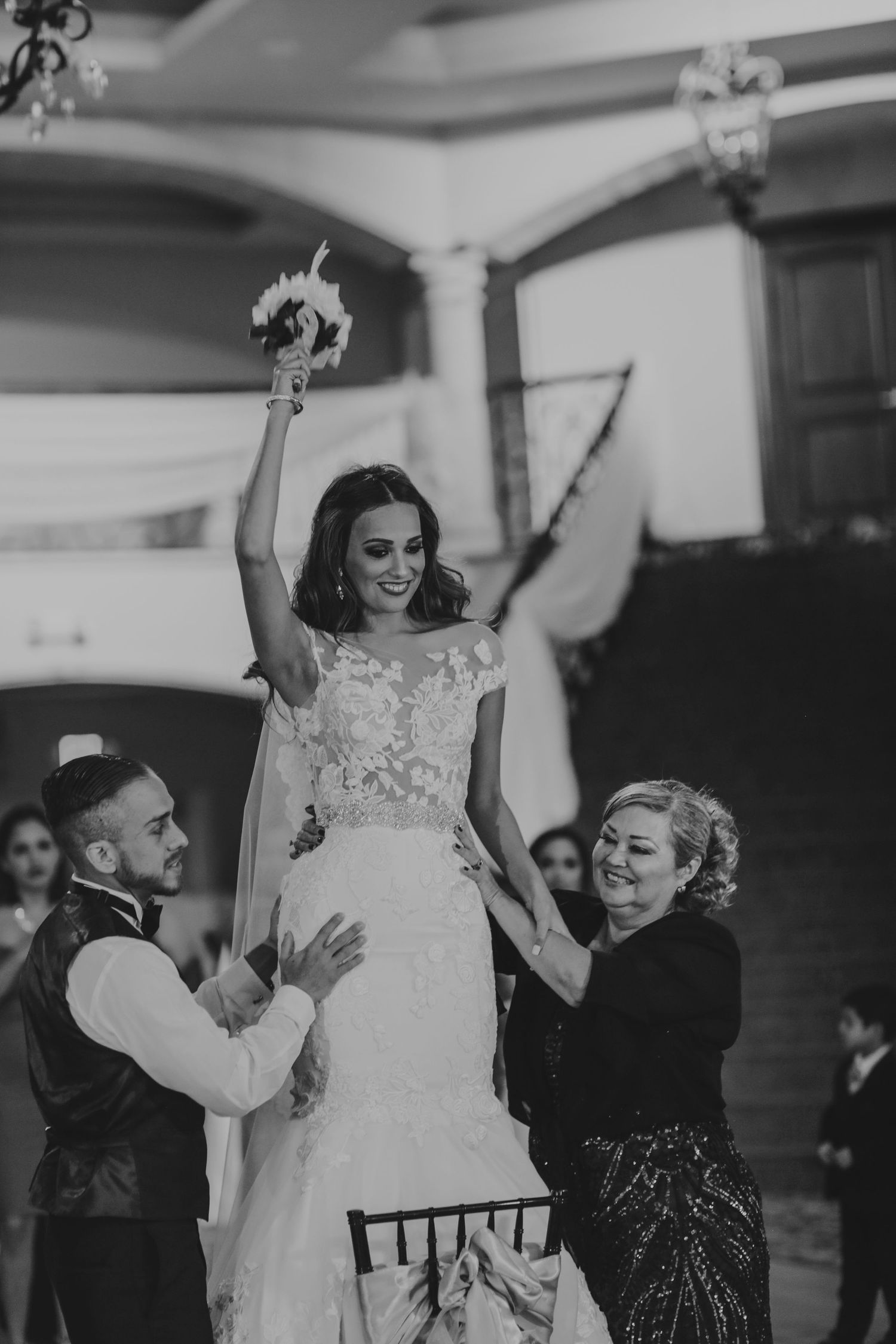 Bride lifted on chair holding bouquet aloft at wedding reception in black and white photo.