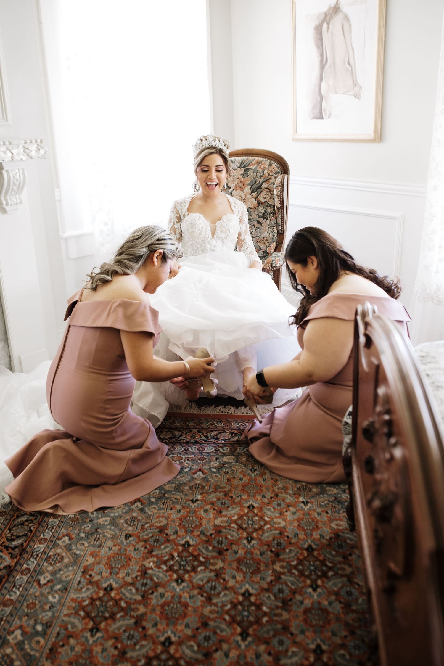 Two bridesmaids in mauve dresses kneel to help bride with shoes in elegant white room.
