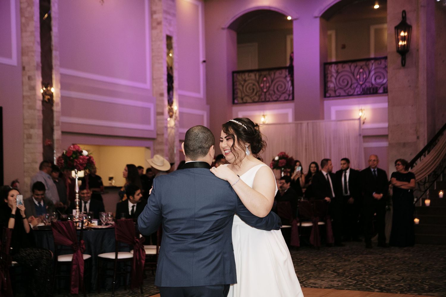 Bride and groom share their first dance in an elegant ballroom with purple uplighting and guests watching.