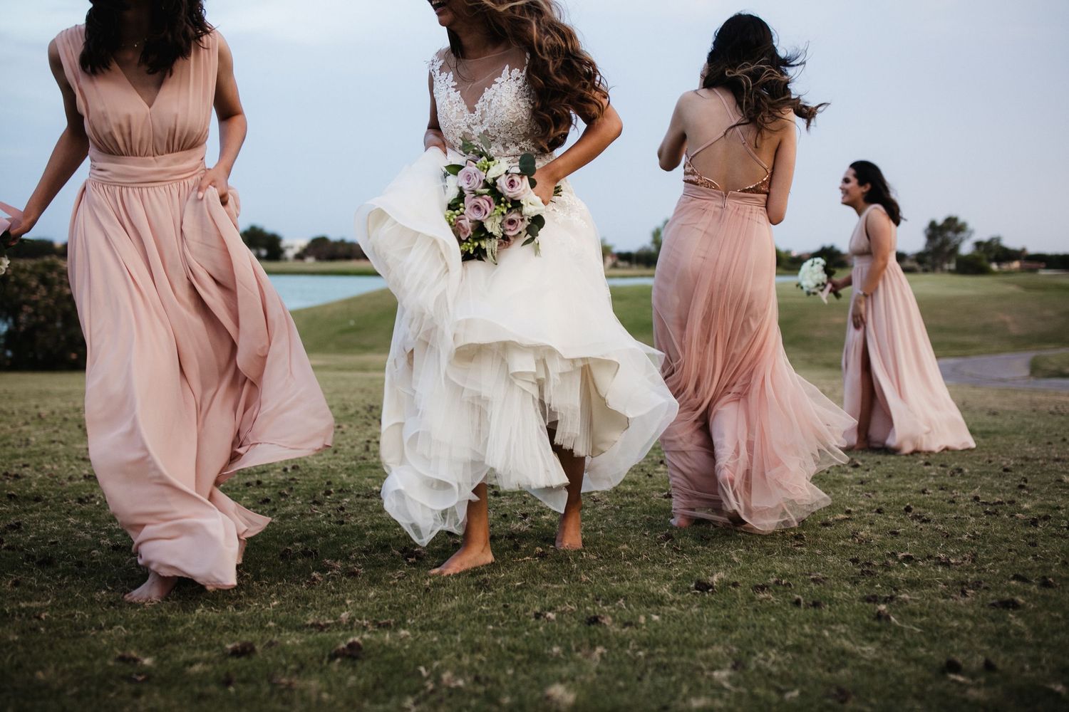 Bride and bridesmaids in blush pink gowns walk barefoot across green grass holding bouquets.