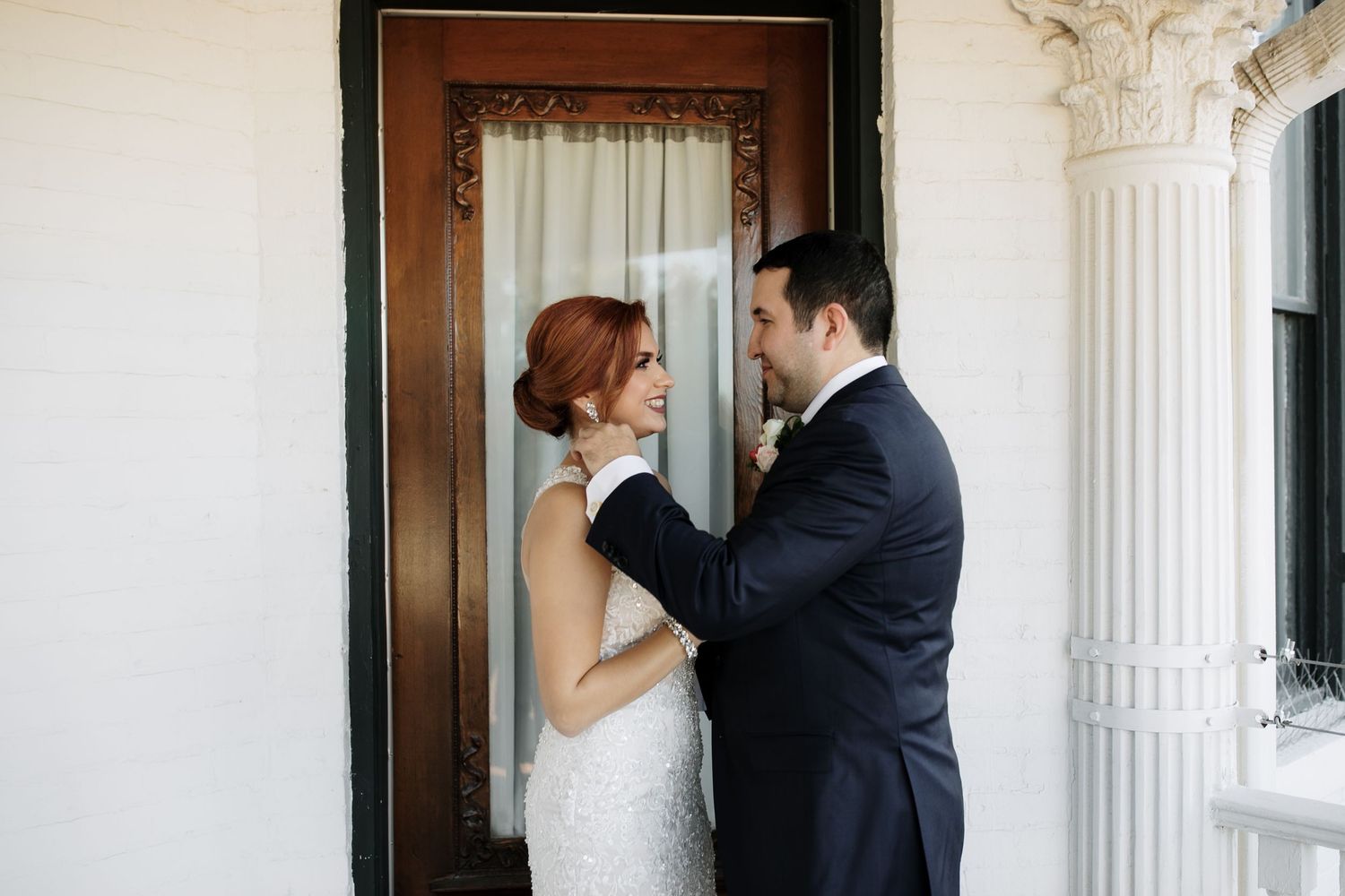 A bride and groom share a tender moment on a porch, with the groom adjusting the bride's hair before their ceremony.