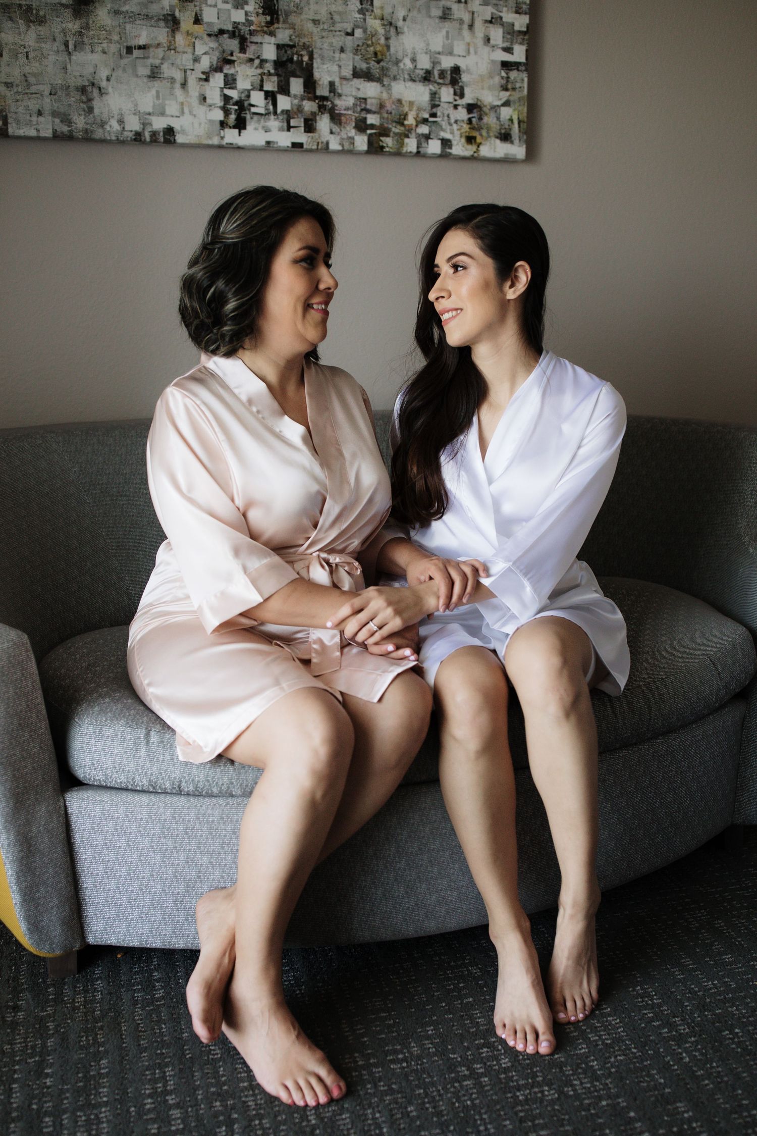 Two women in silk robes sit together on a couch smiling at each other before a wedding.