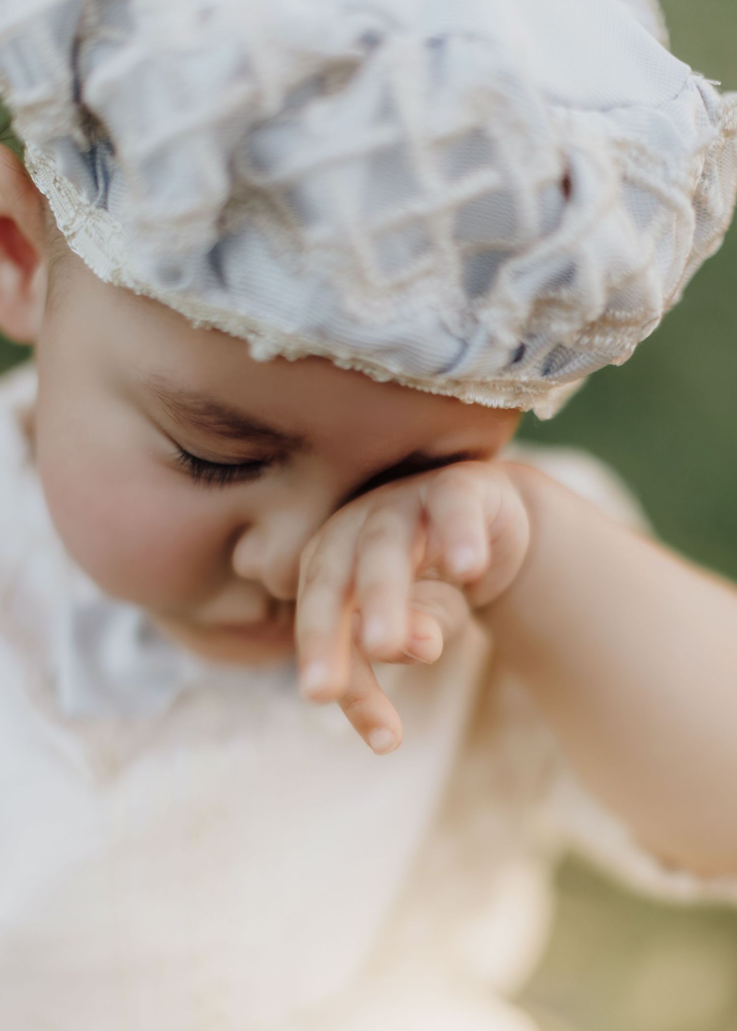 Close up of a baby wearing a white bonnet touching their face.