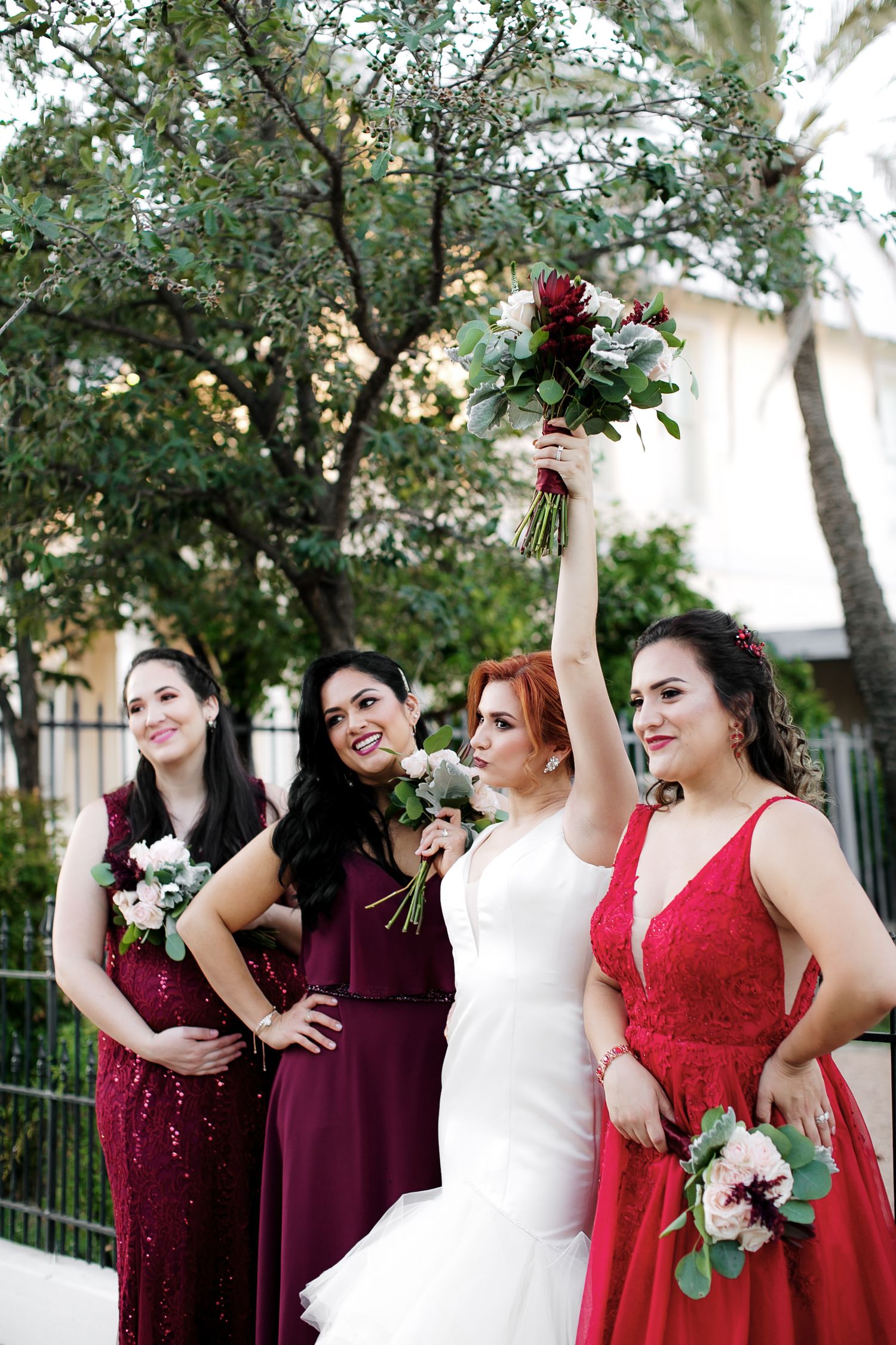 Bride in white gown raises bouquet with three bridesmaids in red and burgundy dresses outdoors.
