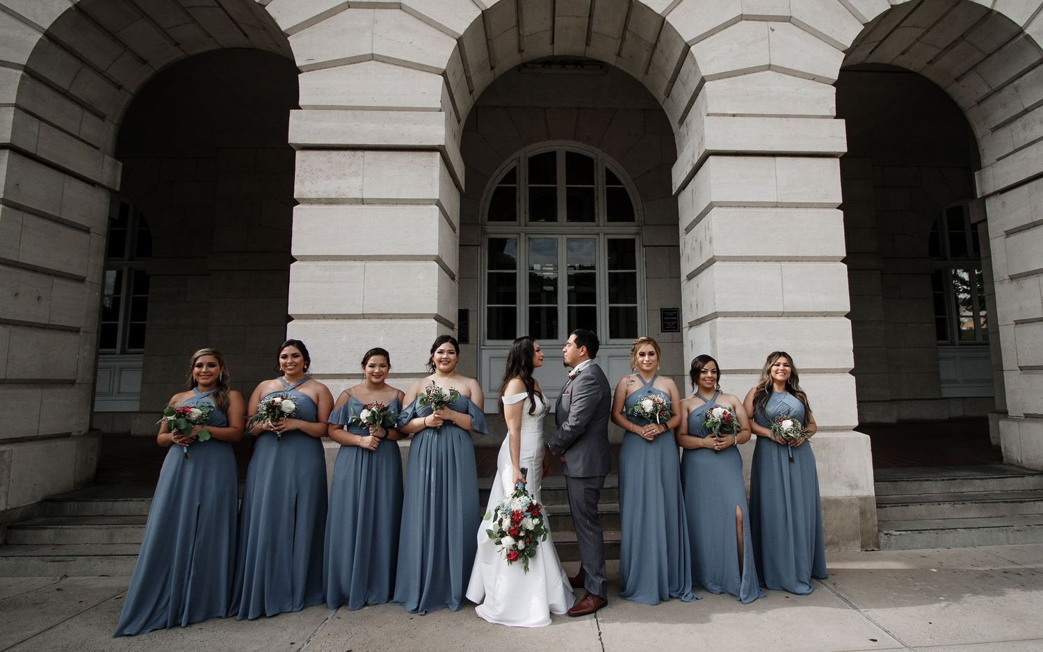 Bride and groom pose with bridesmaids in dusty blue gowns outside grand stone building columns.