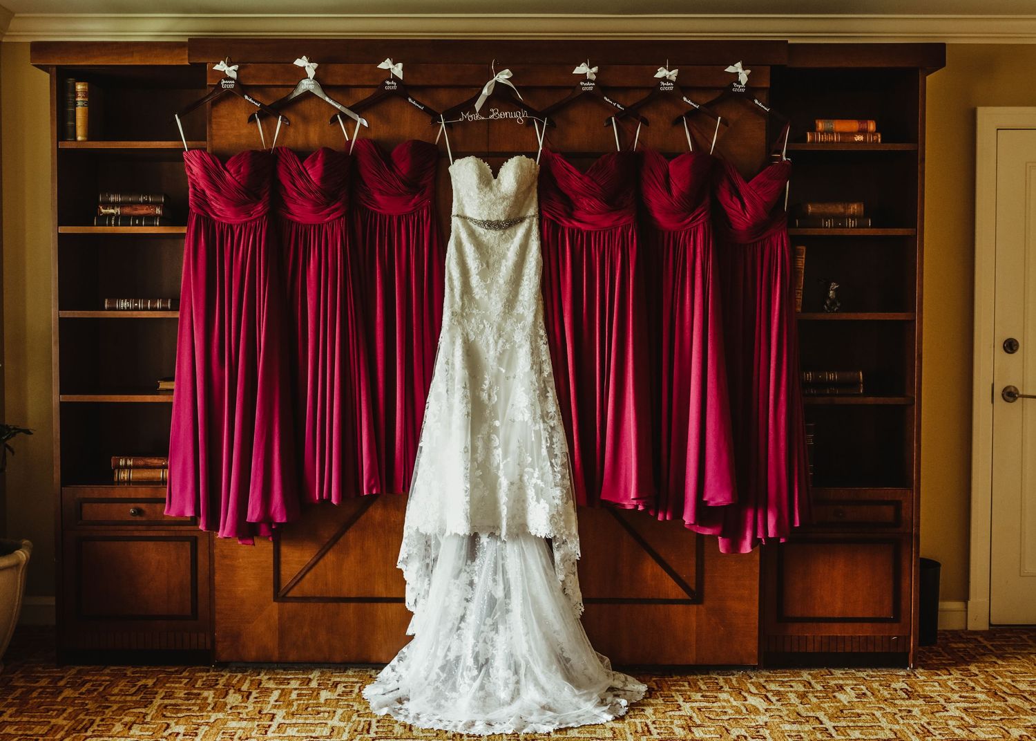 Lace wedding gown hangs beside four burgundy bridesmaid dresses on a wooden bookshelf display.