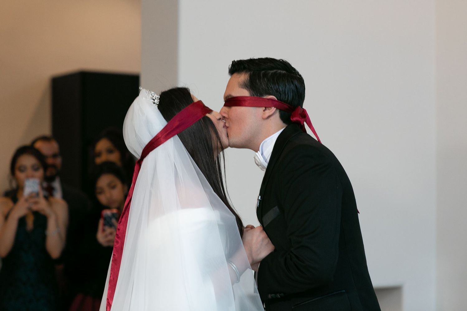 Bride and groom share first kiss at wedding ceremony both wearing red blindfolds as guests watch and photograph.