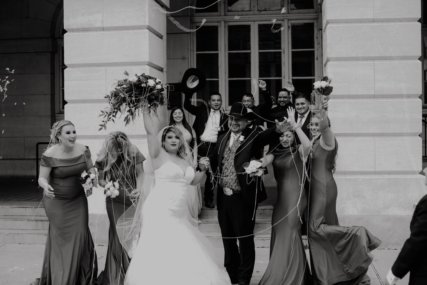 Wedding party celebrating joyfully outside venue in black and white photo.