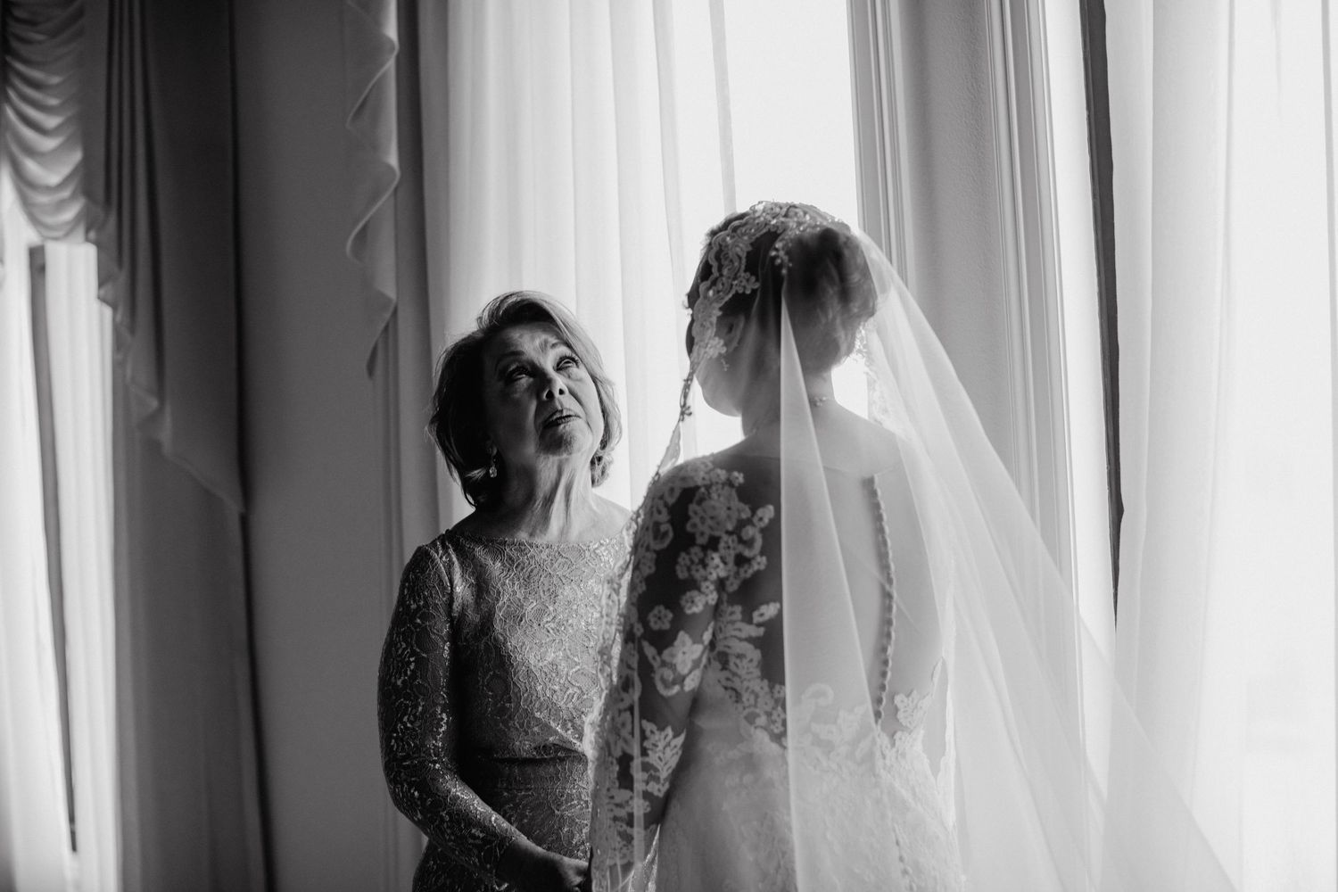Black and white photo of bride in lace veil sharing emotional moment with mother by window.