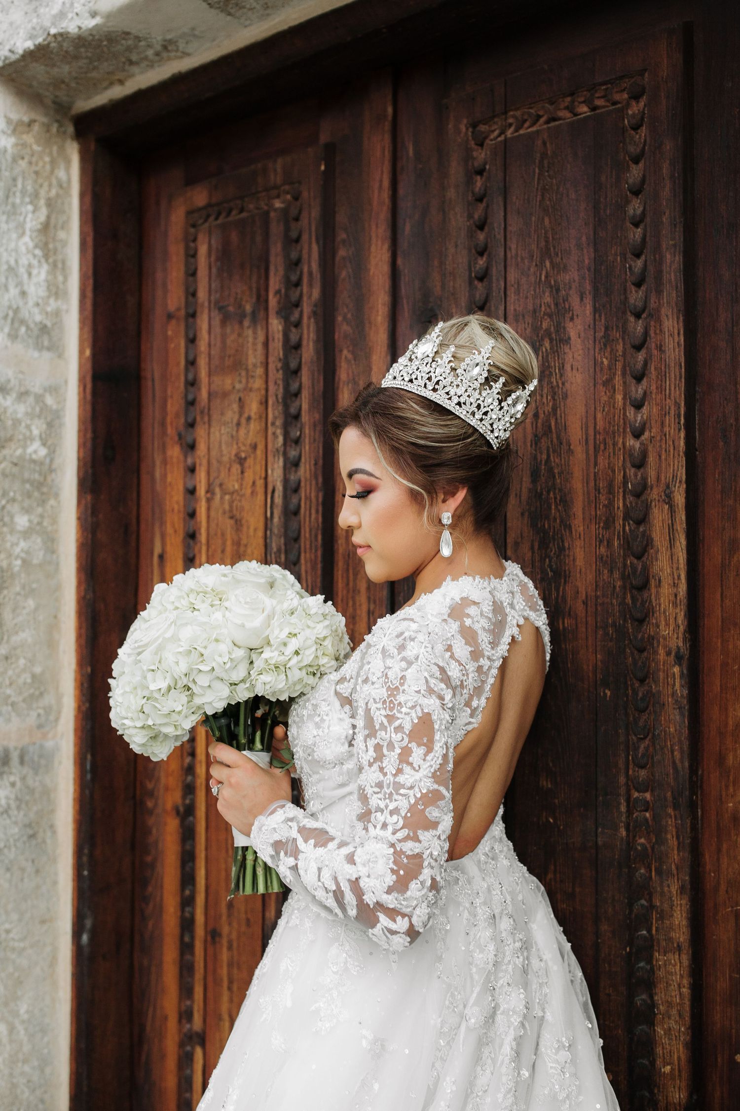 Elegant bride in lace gown with crown tiara holds white hydrangea bouquet before ornate wooden doors.