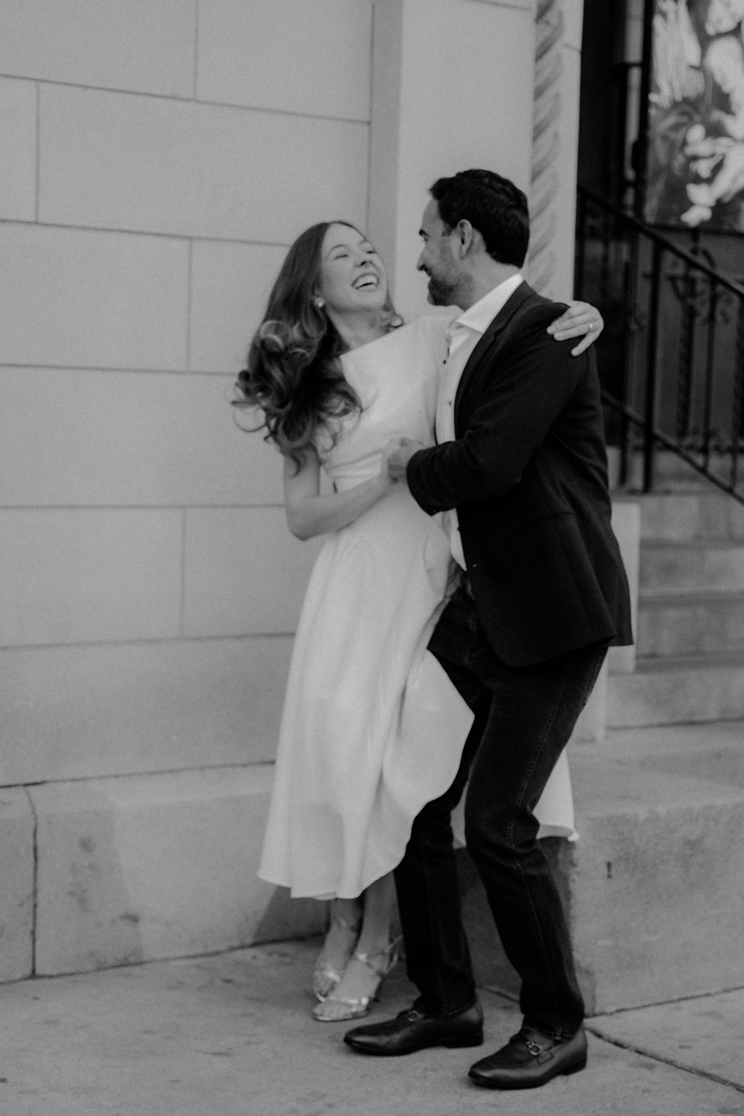 Black and white photo of a laughing couple embracing outdoors, woman in white midi dress, man in dark suit.