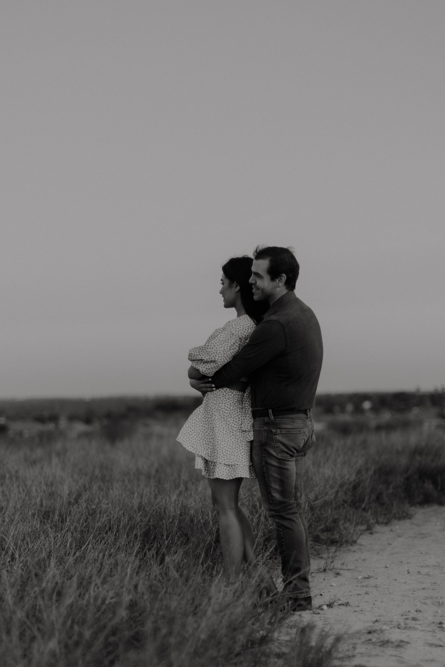 Black and white photo of a couple embracing in a field at dusk, the man holding the woman from behind.