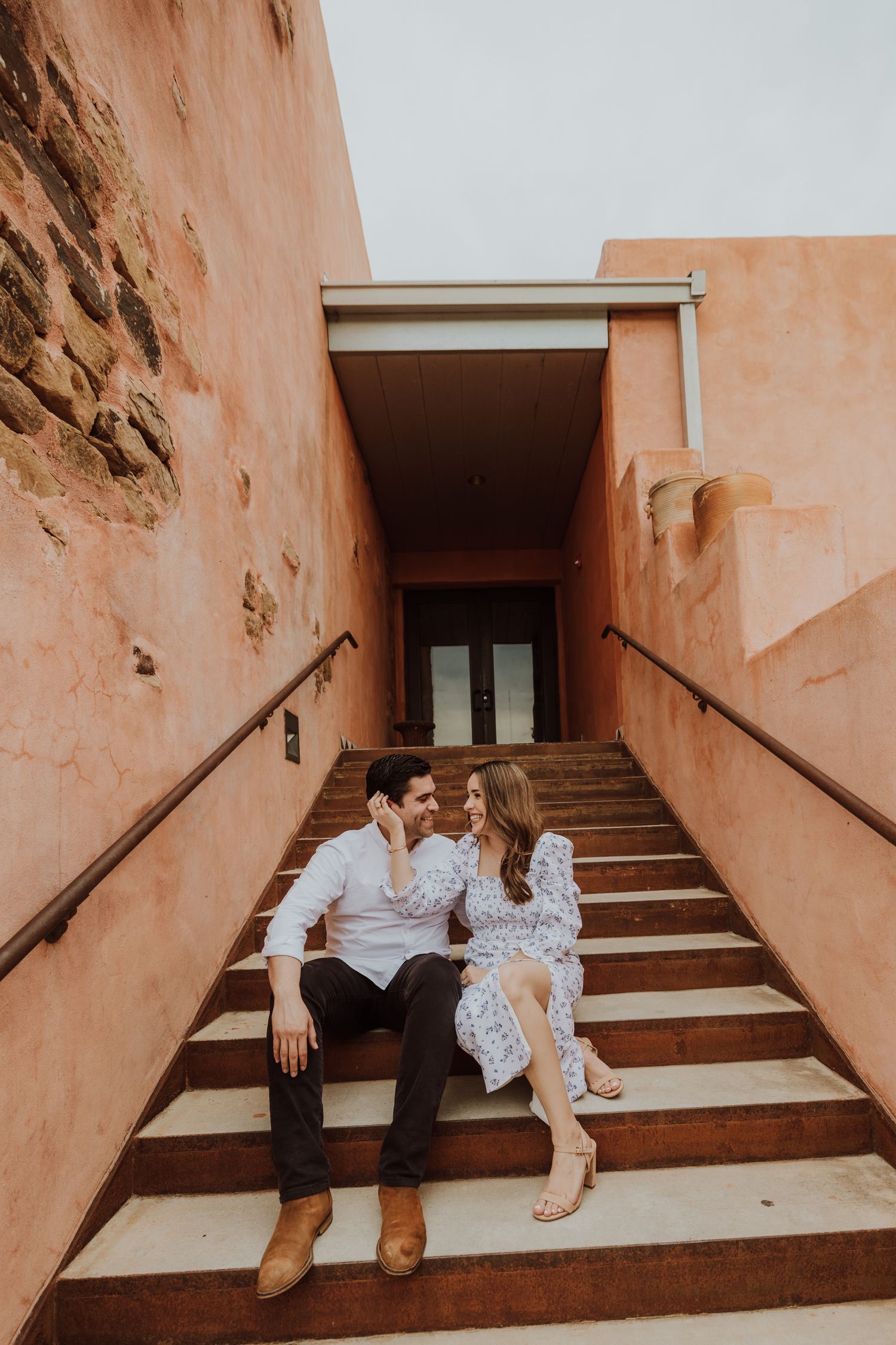Couple sitting on adobe-style staircase, smiling at each other, surrounded by warm terracotta walls.