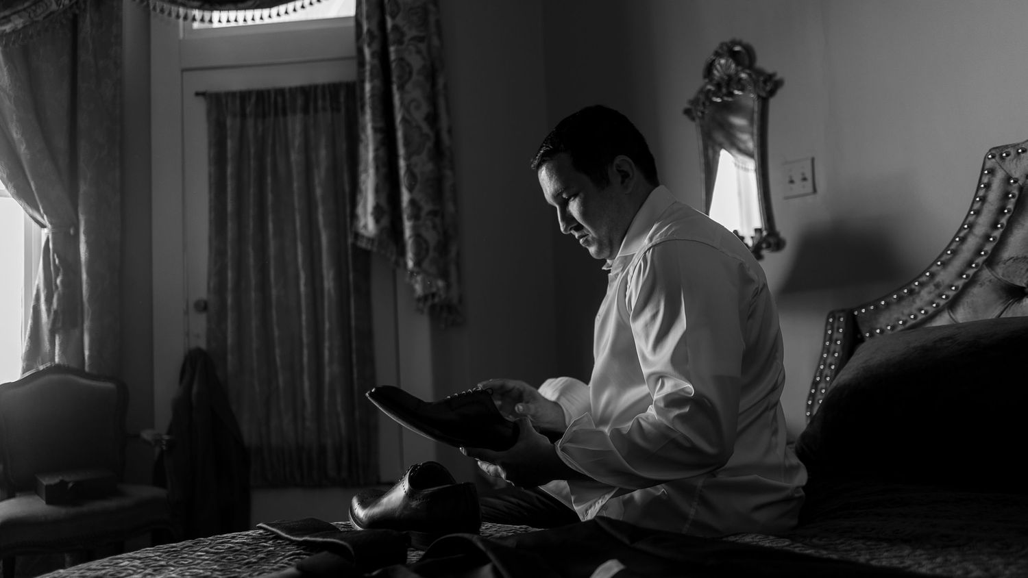 Man in white shirt reads book while sitting on bed in black and white photo.