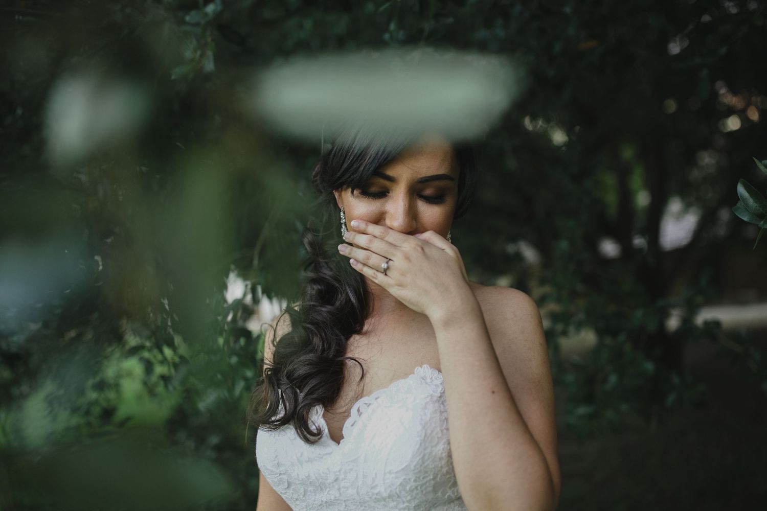 Emotional bride in white lace gown covering face with hand, surrounded by lush green foliage.