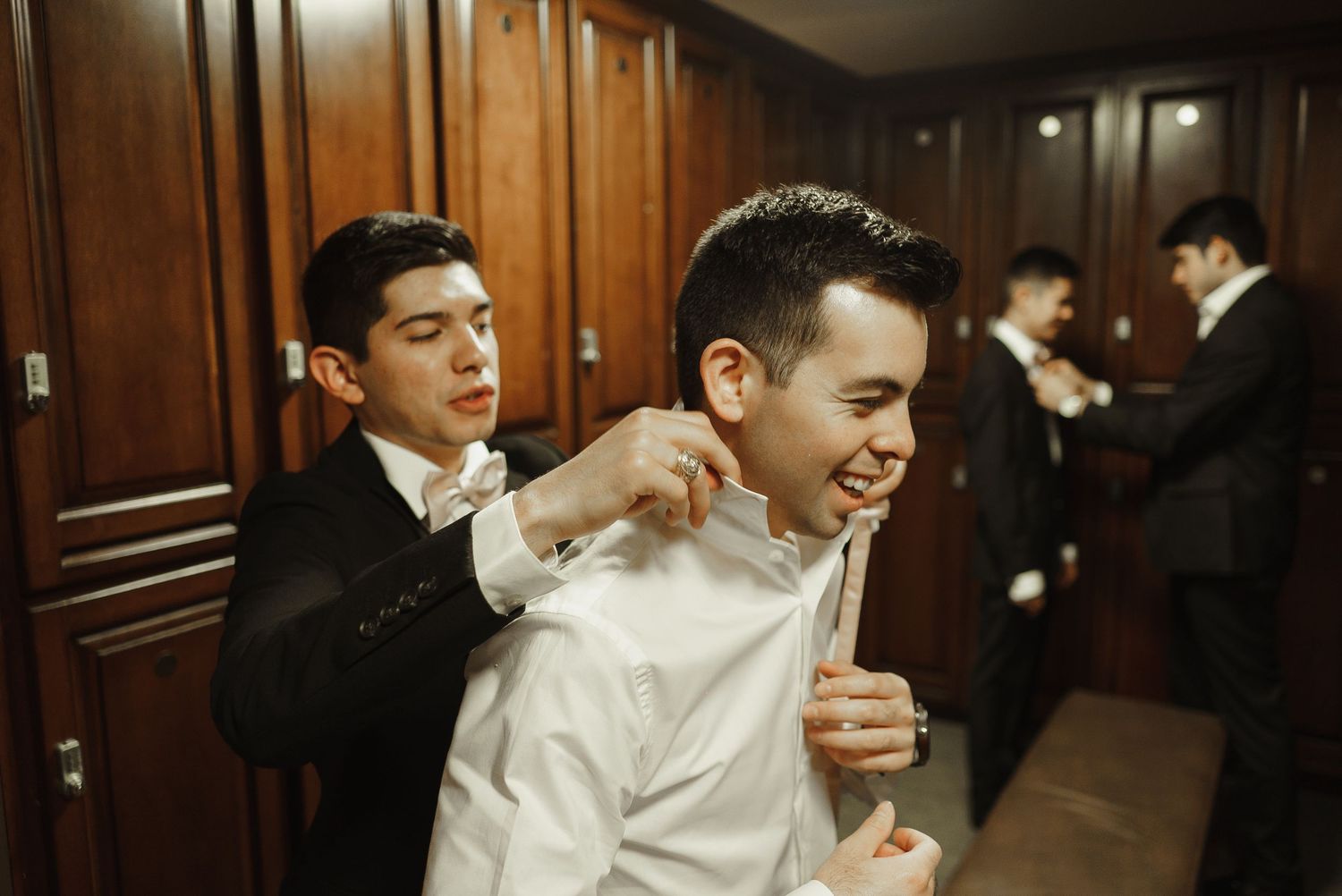 Groomsman helps groom adjust collar in wood-paneled dressing room before wedding ceremony.