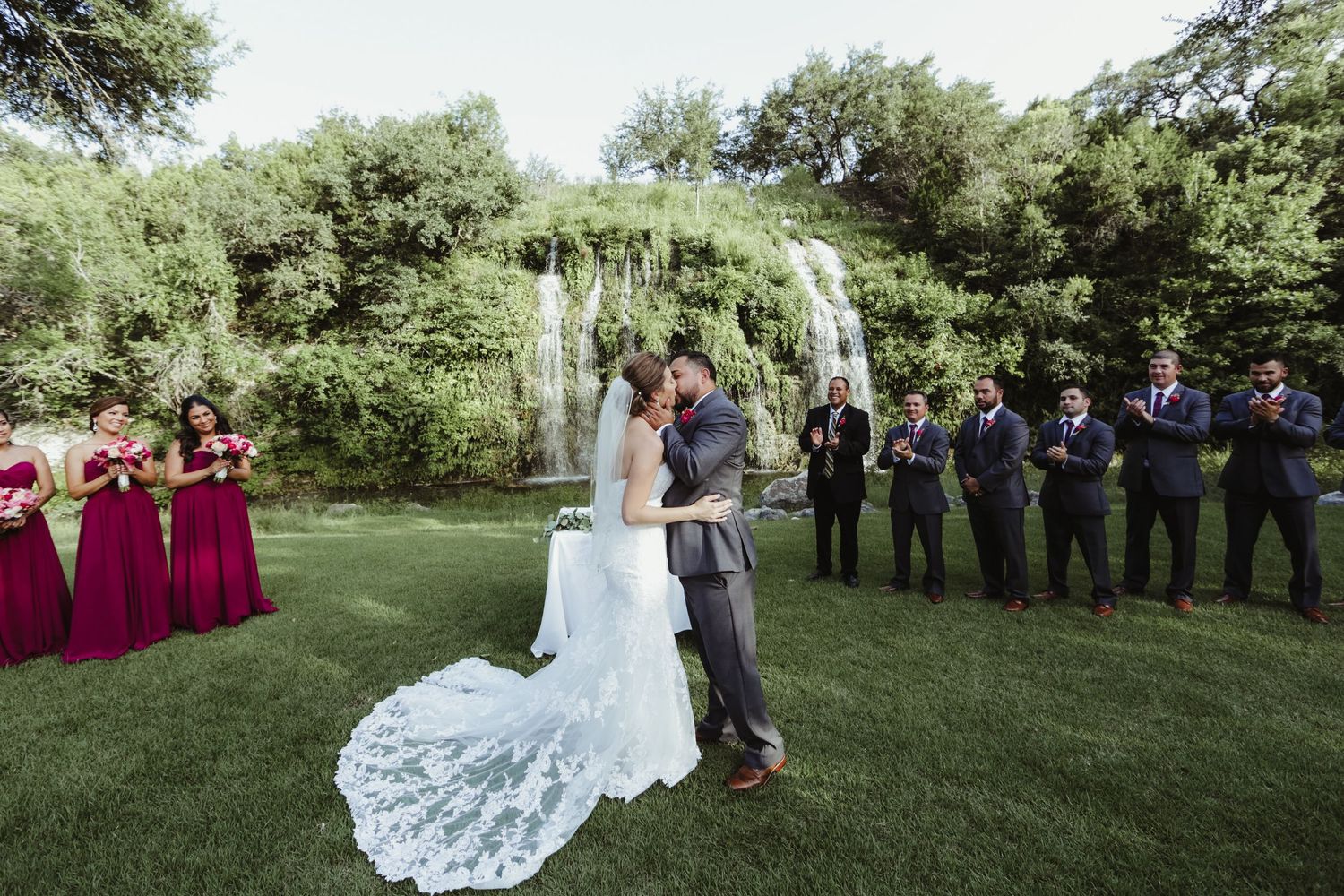 Bride and groom share first kiss at outdoor wedding ceremony with waterfall and lush greenery backdrop.